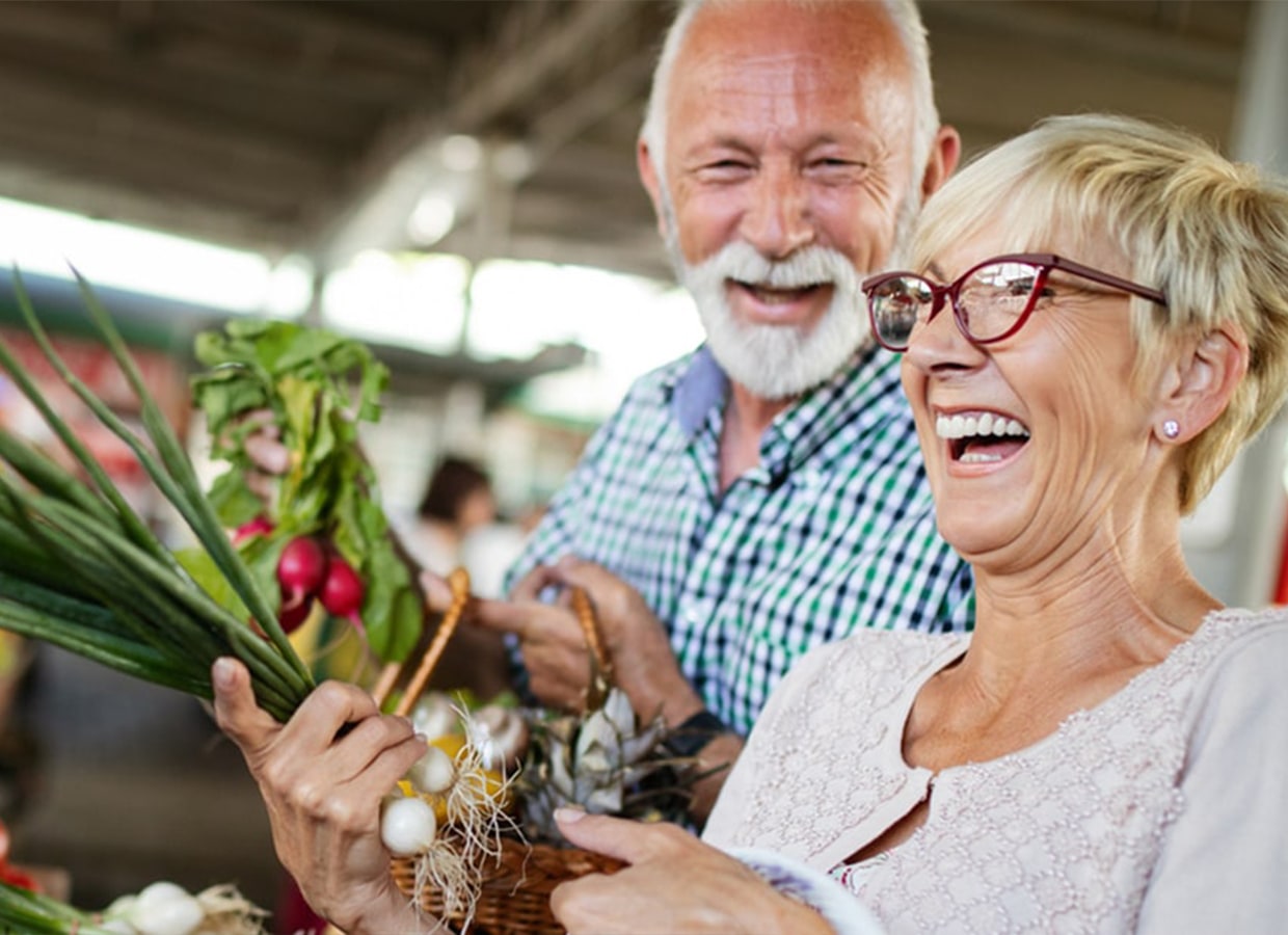 An older man and woman smile and laugh while shopping at a market, holding fresh vegetables and a basket filled with produce. The atmosphere is lively and cheerful.