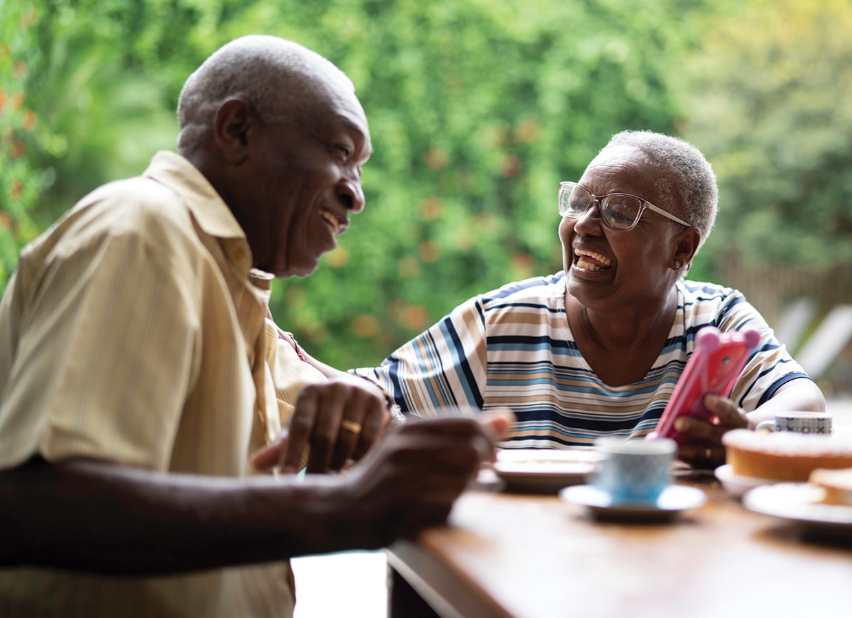 An elderly couple sits at a table outdoors, smiling and laughing together. The woman holds a pink smartphone, and both appear to be enjoying a pleasant conversation, with greenery blurred in the background.