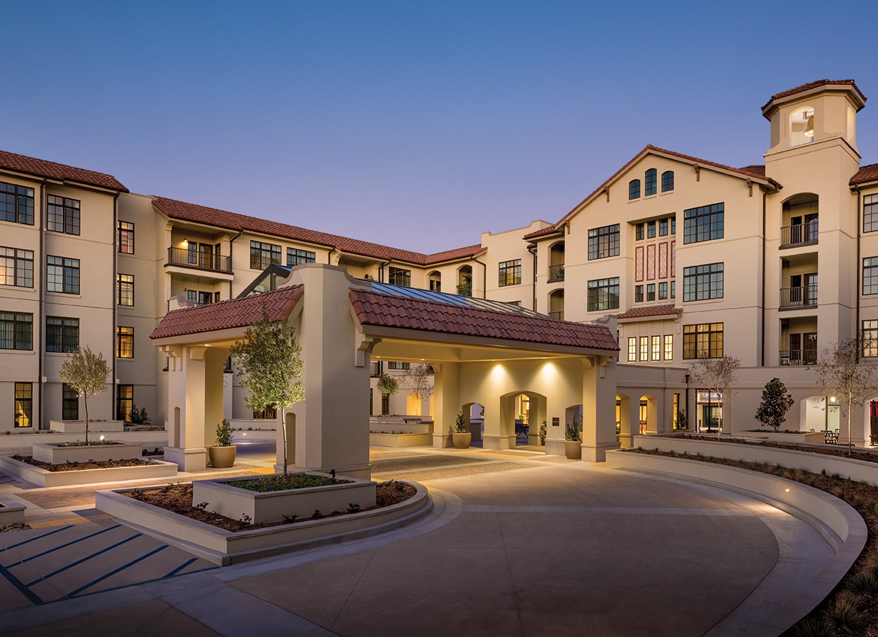 A modern multi-story building with cream-colored walls, red-tiled roofs, large windows, and a covered entrance, surrounded by landscaped pathways and small trees, photographed at dusk with exterior lights on.