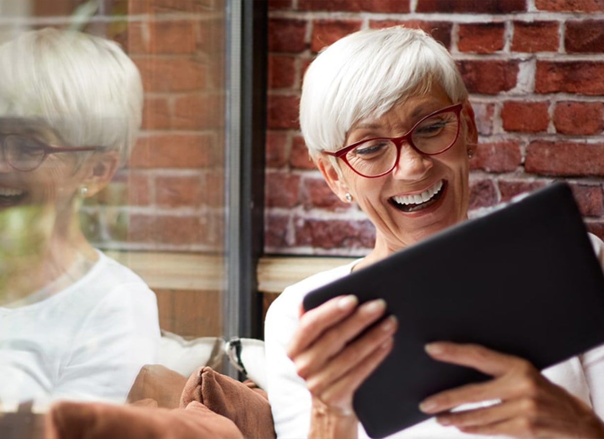 An older woman with short white hair and red glasses is smiling and holding a tablet. She is sitting by a window with a brick wall behind her, and her reflection is visible in the glass.