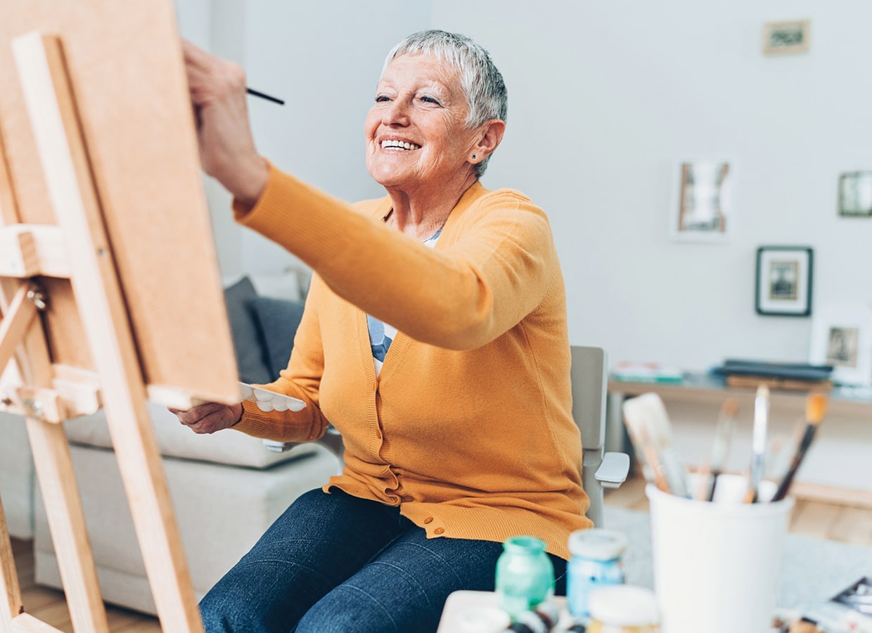 An older woman with short gray hair, wearing a yellow cardigan, smiles while painting at an easel in a bright, cozy living room. Art supplies and brushes are visible in the foreground.