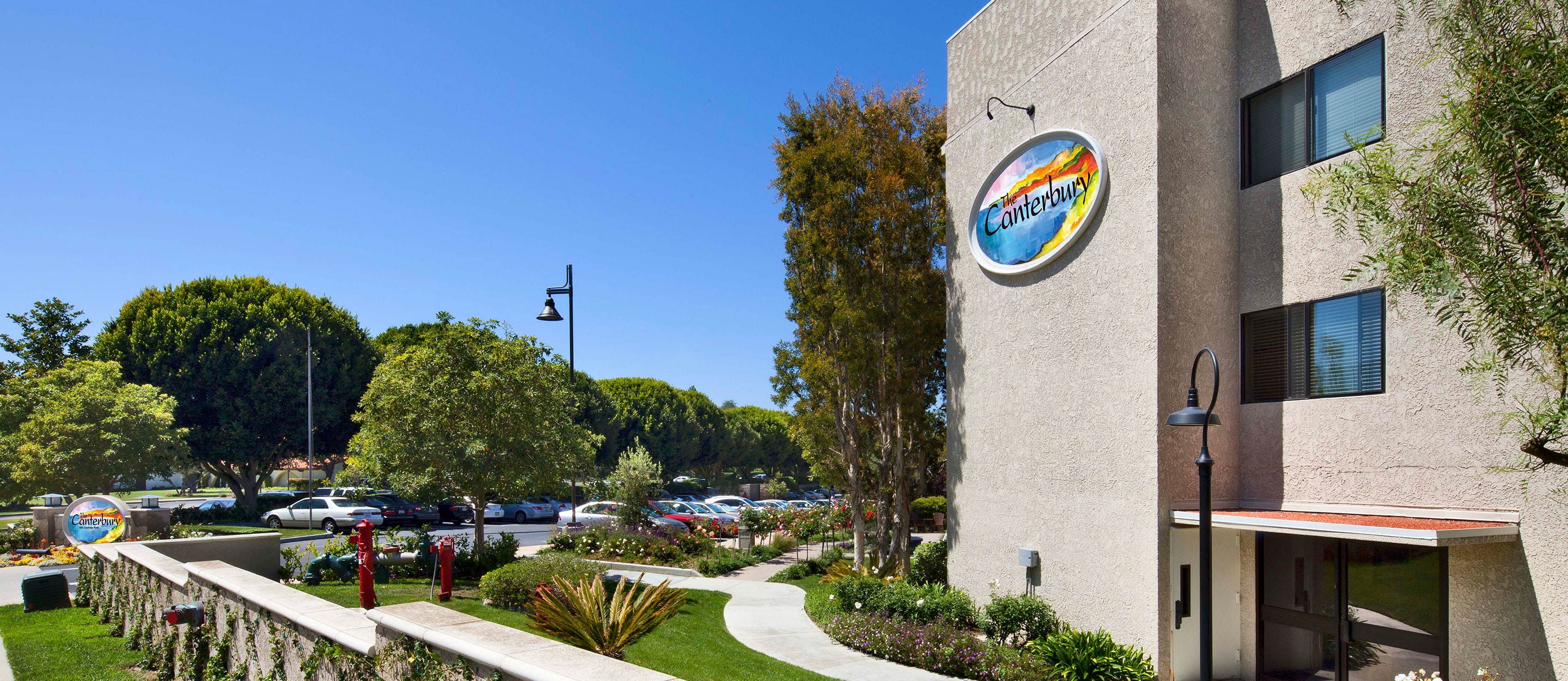 A sunny day outside a beige building with a colorful Canterbury sign, surrounded by landscaped gardens, trees, a walkway, and a parking lot with several parked cars.