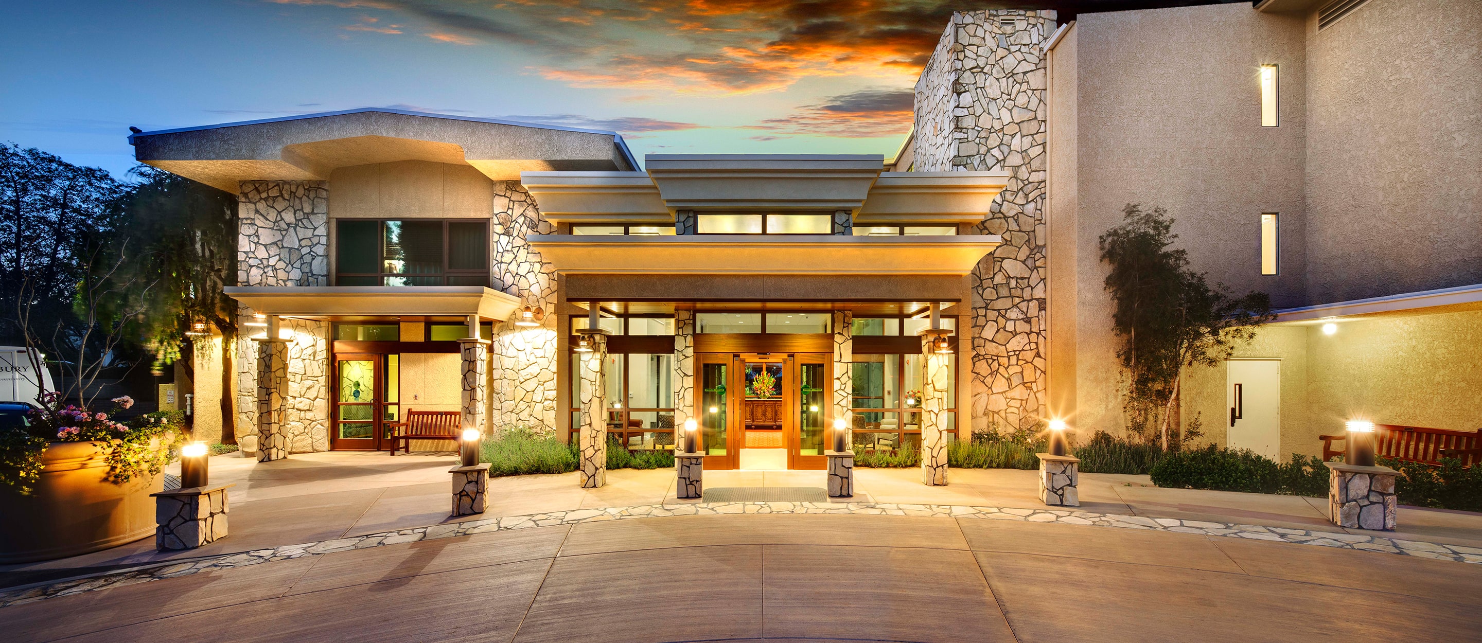 The image shows the well-lit entrance of a modern building with large windows, stone walls, and decorative lamps, set against a vibrant sunset sky. The driveway curves toward the main doors, creating a welcoming atmosphere.