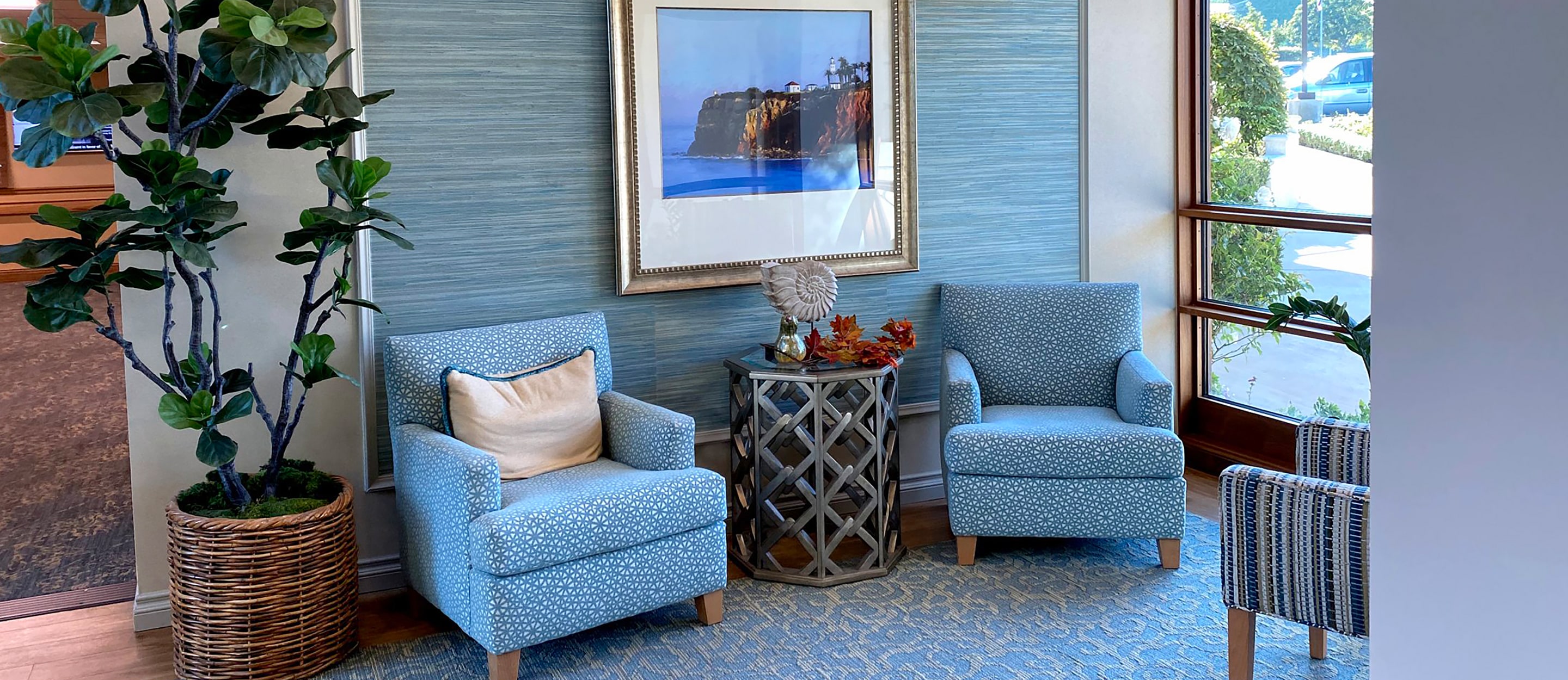 Two light blue armchairs with a pillow sit beside a small round table with a decorative item and autumn leaves. A potted plant and framed beach photo are behind them. Sunlight streams through large windows on the right.