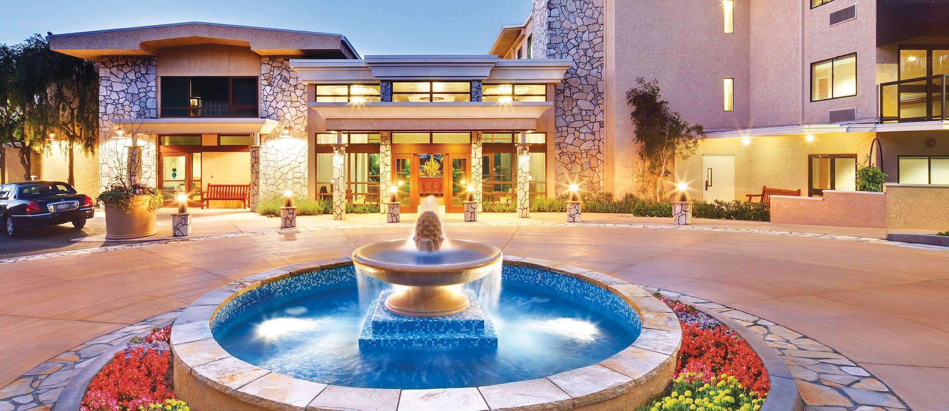 A modern building with stone accents and large windows features a circular fountain in front, surrounded by colorful flower beds, at dusk with soft exterior lighting.