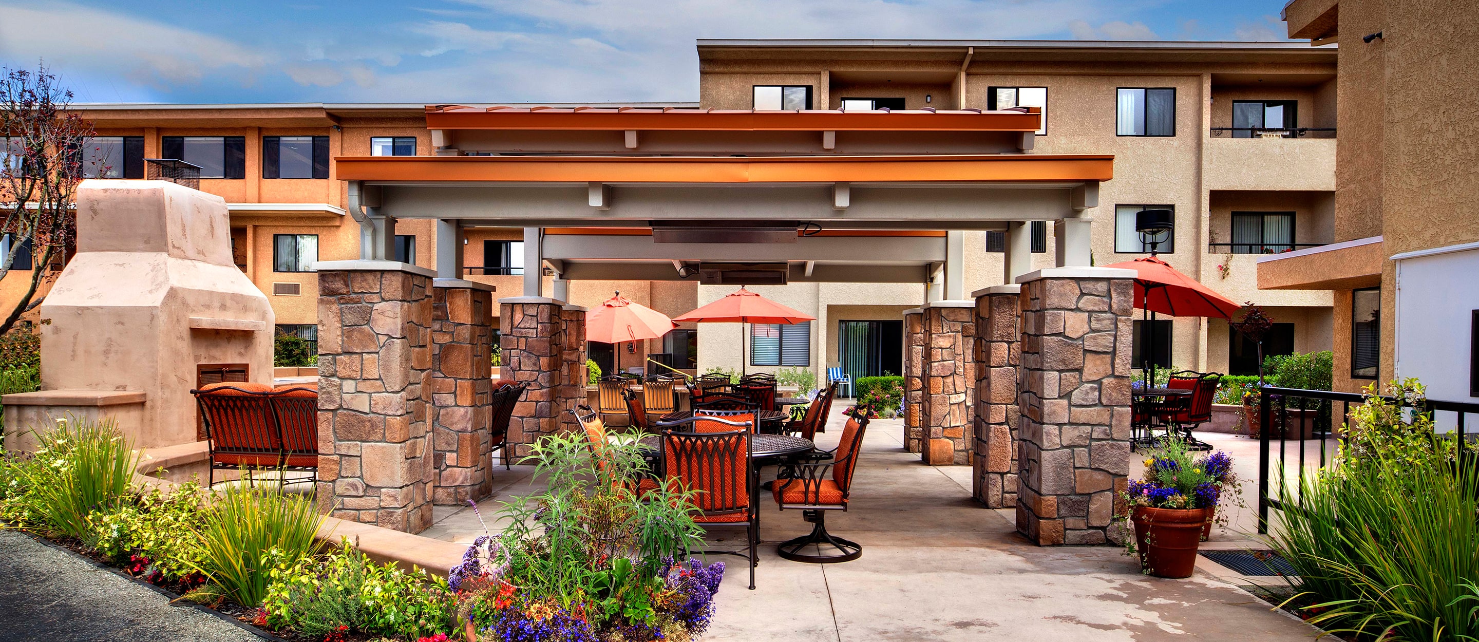 A sunny outdoor patio area with tables, chairs, and red umbrellas, surrounded by landscaped plants and flowers, next to a multi-story building with beige and orange walls.