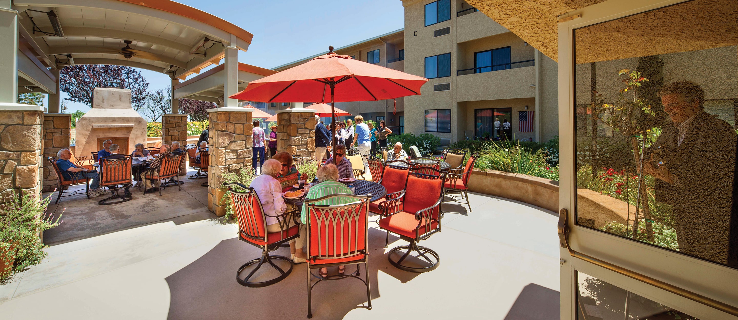 Seniors gather on a sunny patio, sitting at tables with red chairs and umbrellas, socializing and dining near a fireplace; others are mingling in the background, with a reflection of a person in a window.