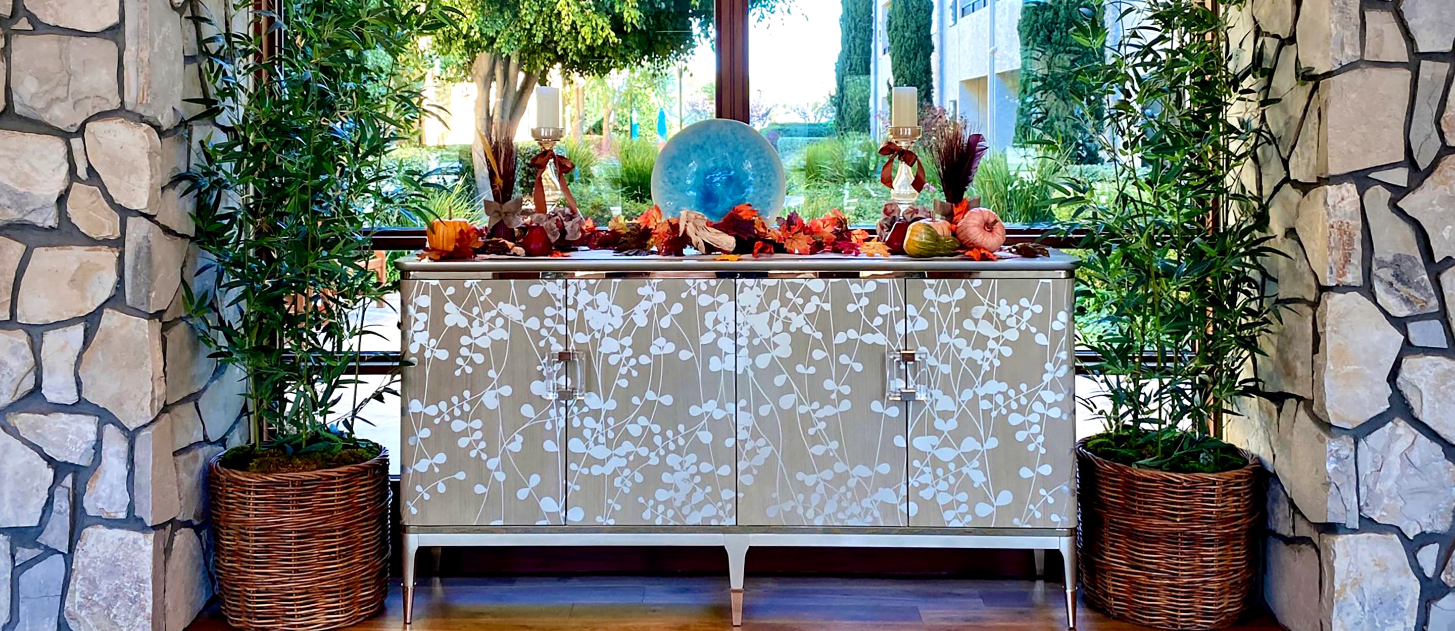 A modern floral-patterned sideboard is decorated with autumn leaves, fruit, and candles. Two large potted plants flank the sideboard, and a blue glass sculpture sits in the center, with trees visible through a large window behind.