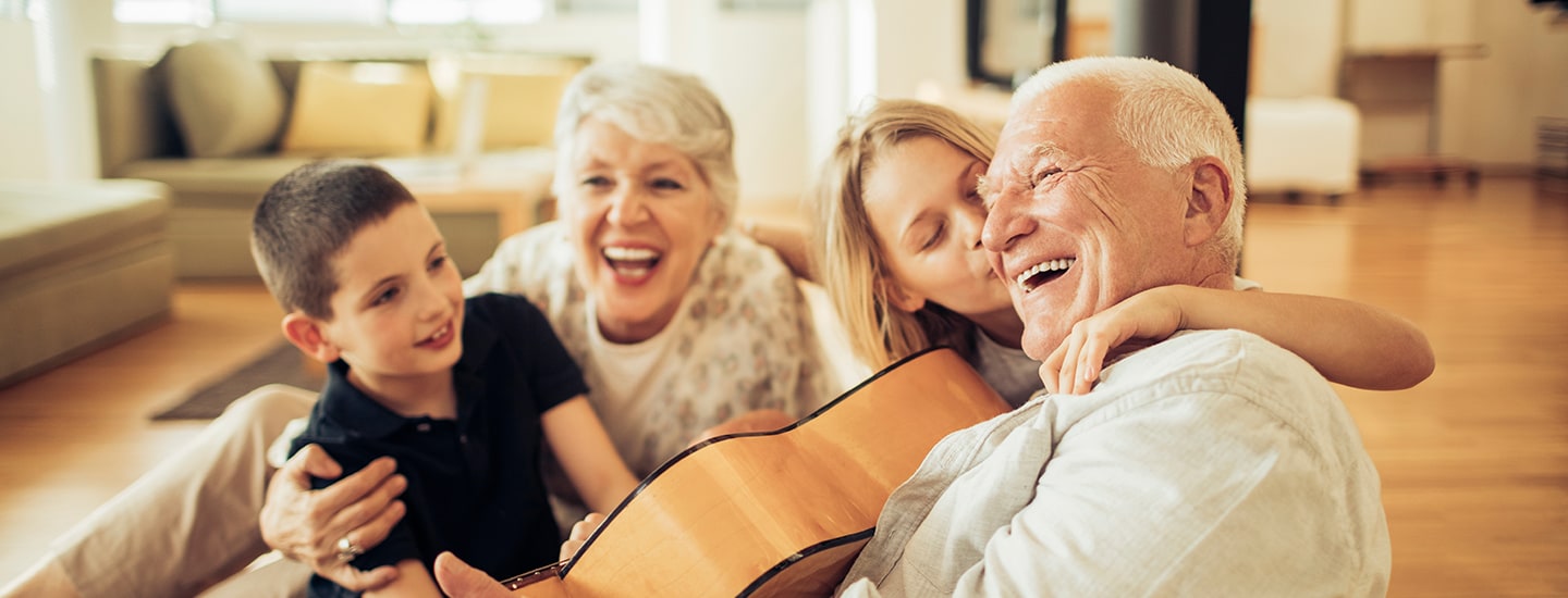 An elderly couple sits on the floor with two children. The man holds a guitar and laughs while a young girl hugs and kisses him on the cheek. The woman and boy smile joyfully in a brightly lit living room.