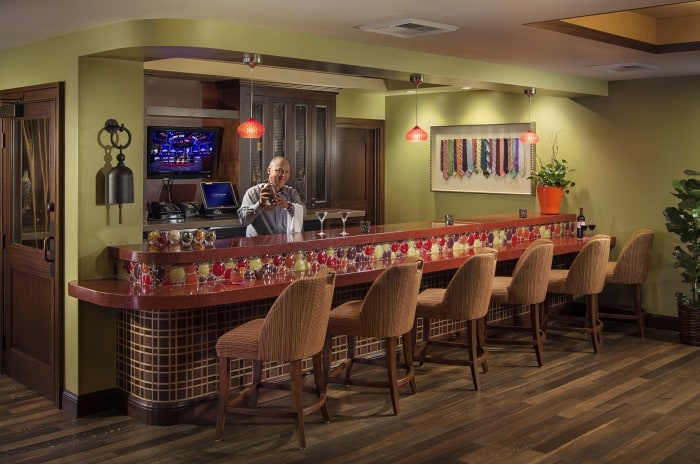 A bartender stands behind a colorful, modern bar with red accents and cushioned chairs. Glasses and bottles are on the counter, with a TV, artwork, and plant in the background, creating a cozy, inviting atmosphere.