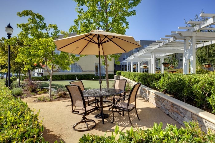 A patio area with a round metal table and four chairs under a large beige umbrella, surrounded by greenery and a bocce ball court, near a pergola and a building in the background on a sunny day.