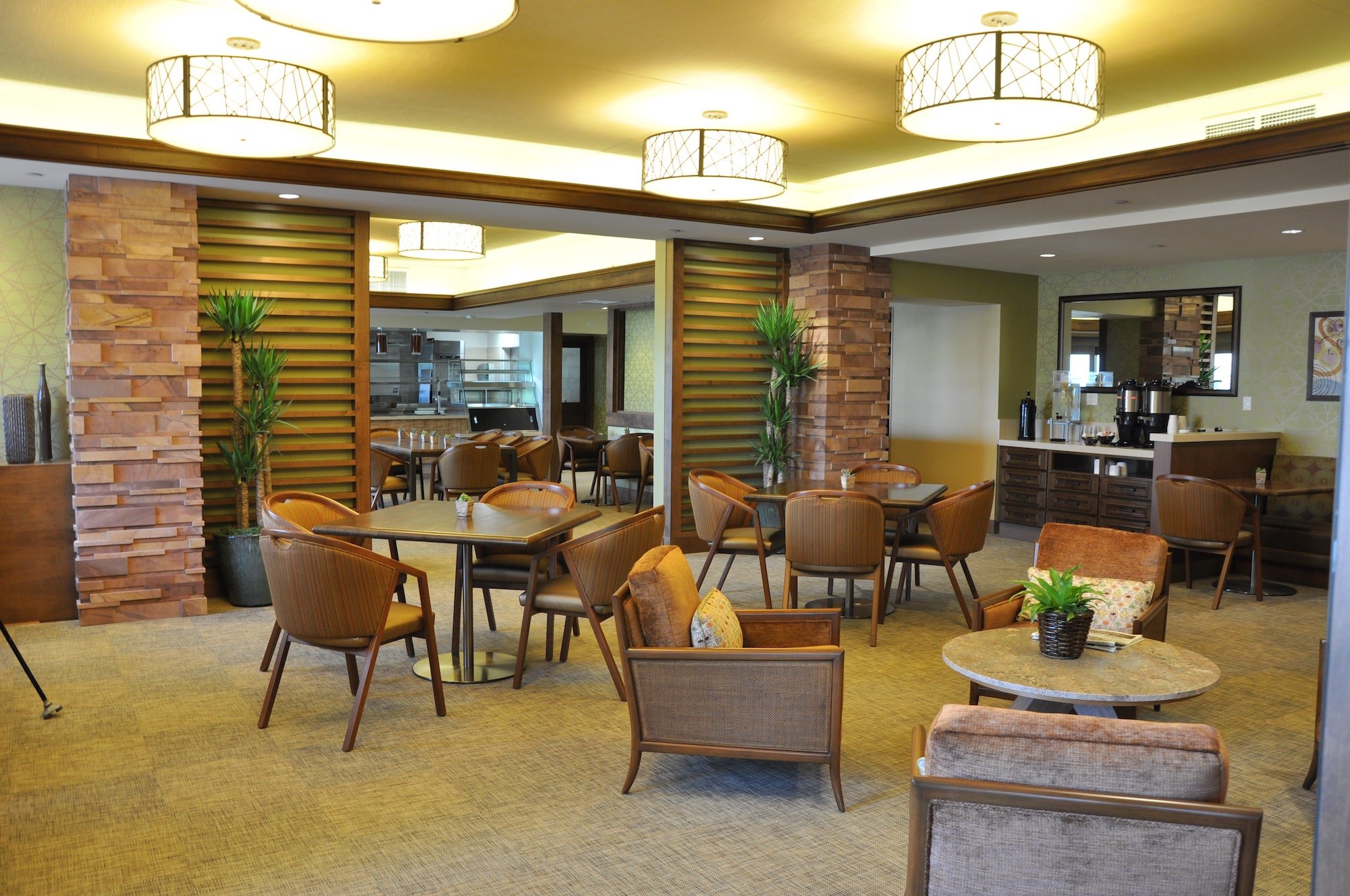 A modern lounge with round tables and wicker chairs, beige carpet, stone accents, potted plants, and warm lighting. There is a coffee station and shelves in the background.