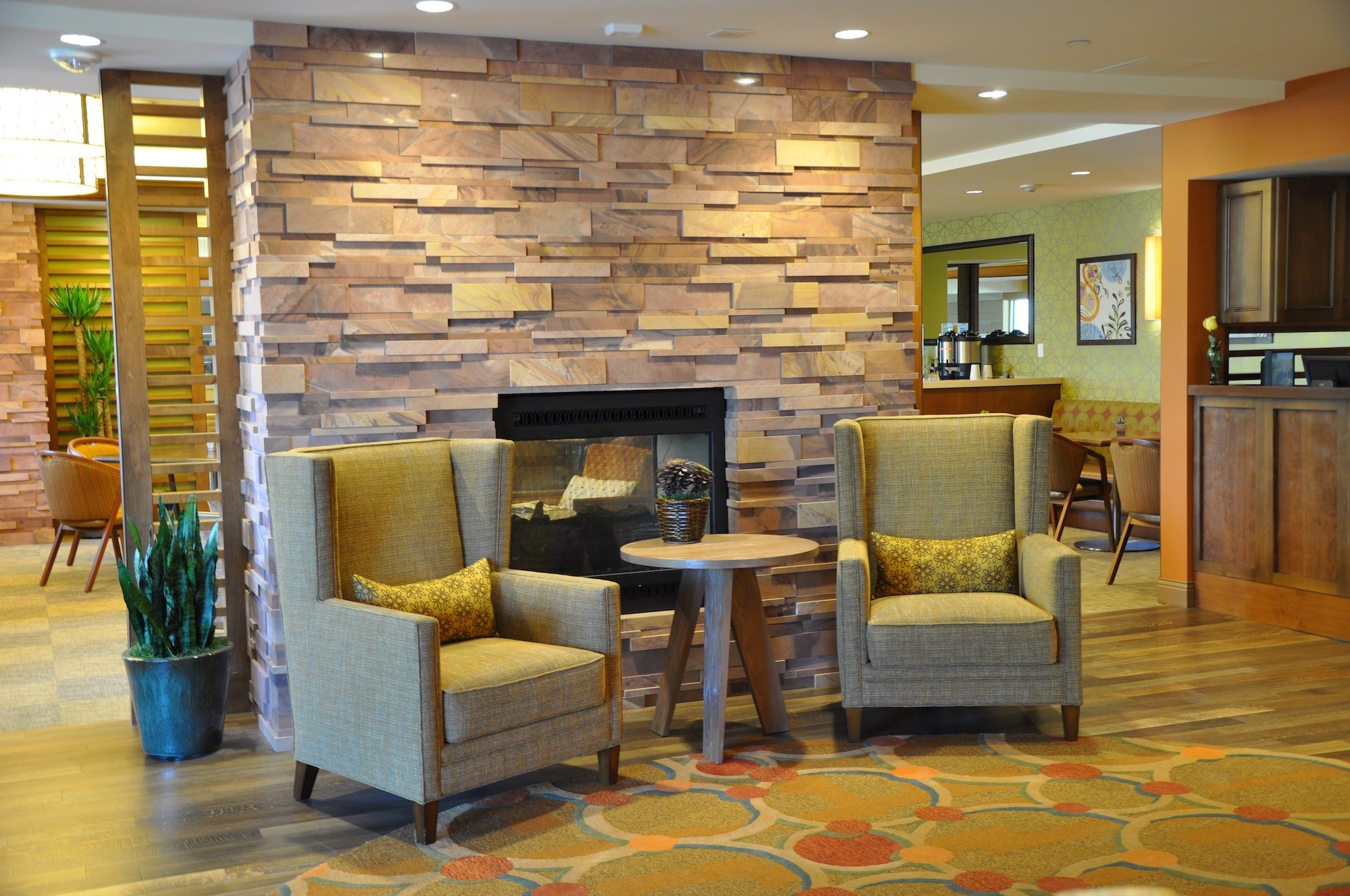 Two armchairs with yellow cushions sit beside a small round table in front of a modern stone fireplace. The area has wood floors, warm lighting, and a decorative plant, creating a cozy, inviting atmosphere.