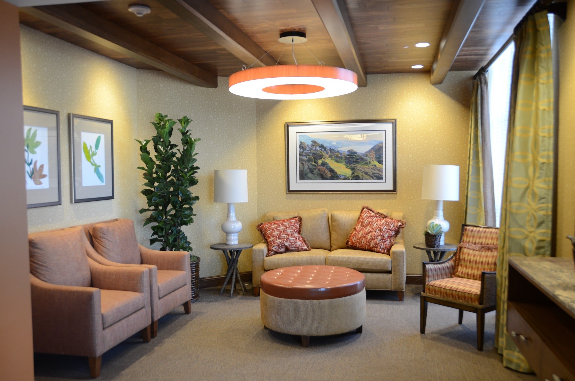 A cozy living room with beige armchairs, a tan sofa with patterned cushions, a round leather ottoman, potted plant, framed art, two table lamps, and a large round ceiling light.