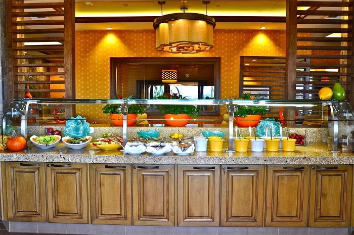 A buffet with various salads, fruits, and bowls displayed on a granite counter. Wooden cabinets are below, and bright orange accent walls and modern lighting decorate the background.