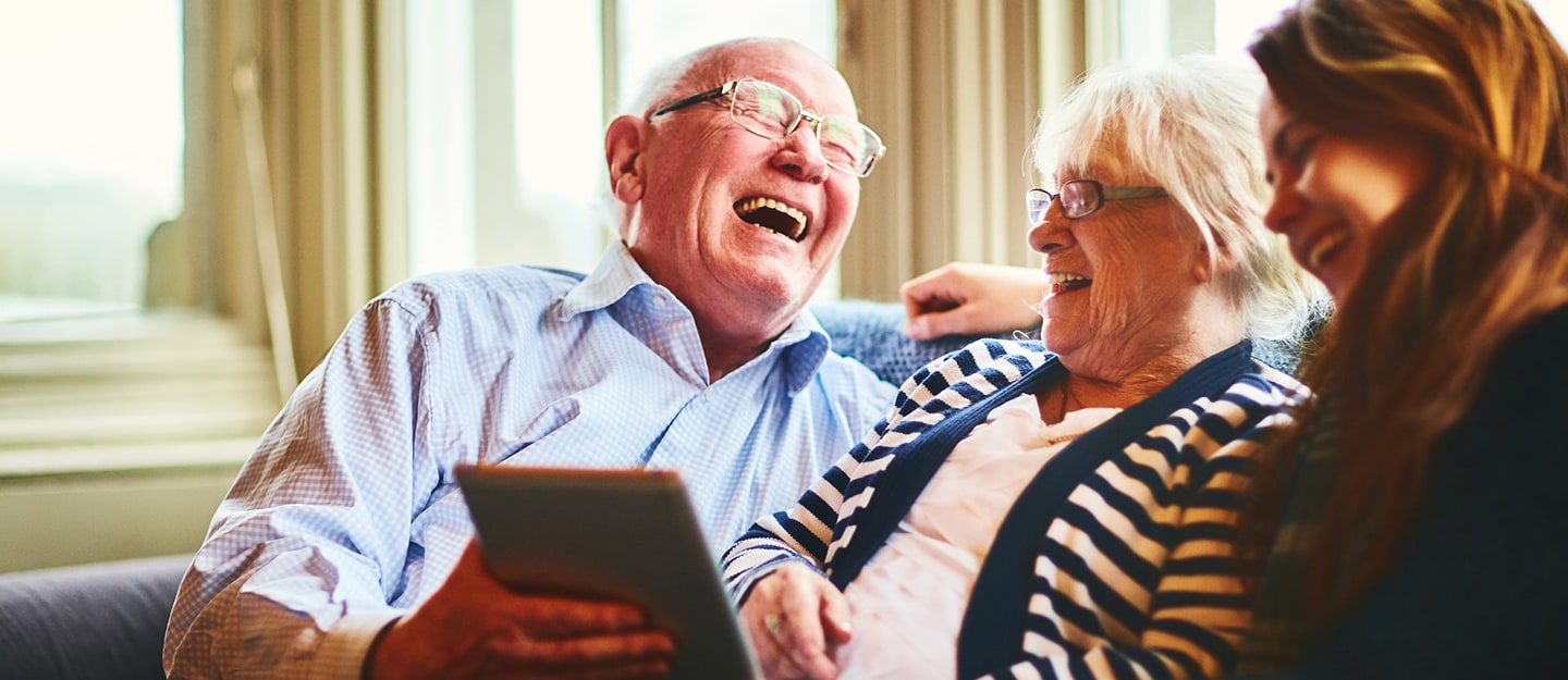 An elderly couple and a young woman sit on a couch, laughing together while holding a tablet in a bright, cozy room with large windows in the background.