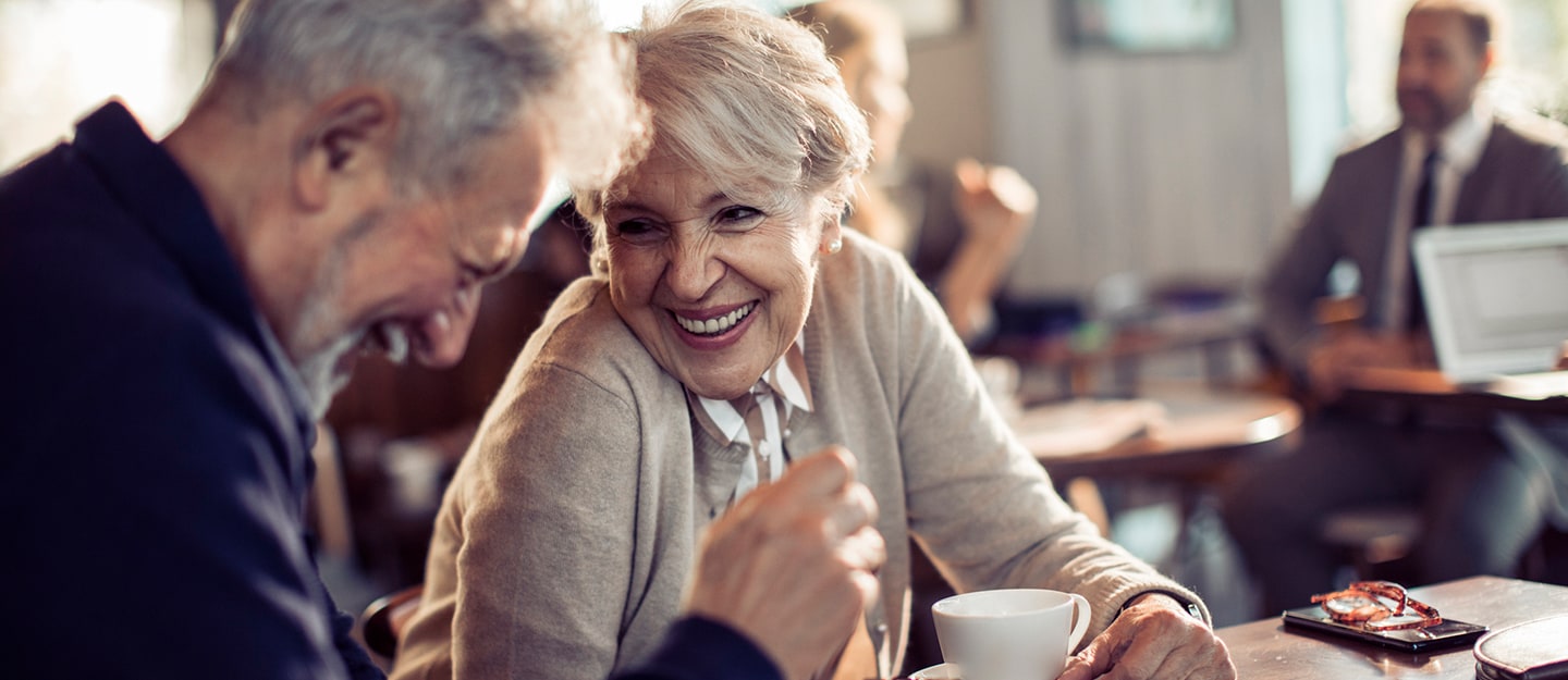 An older man and woman sit at a table in a café, smiling and laughing together over coffee. Other people are visible in the blurred background, creating a warm, social atmosphere.
