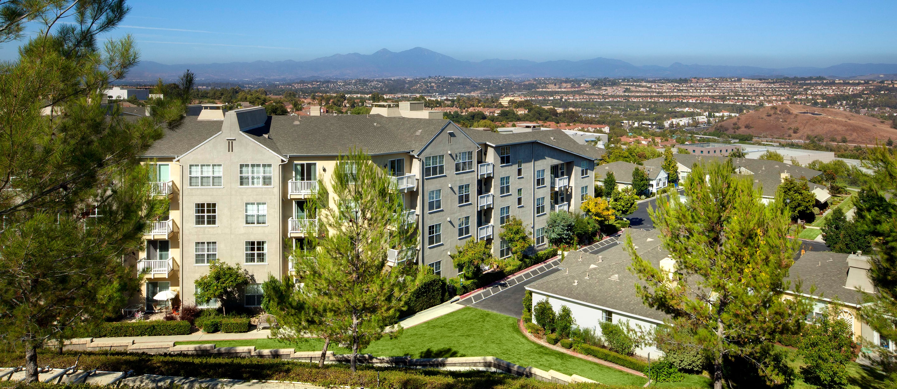 A large apartment complex with gray buildings, balconies, green lawns, and trees in the foreground. In the distance, rolling hills, scattered buildings, and mountains under a clear blue sky.