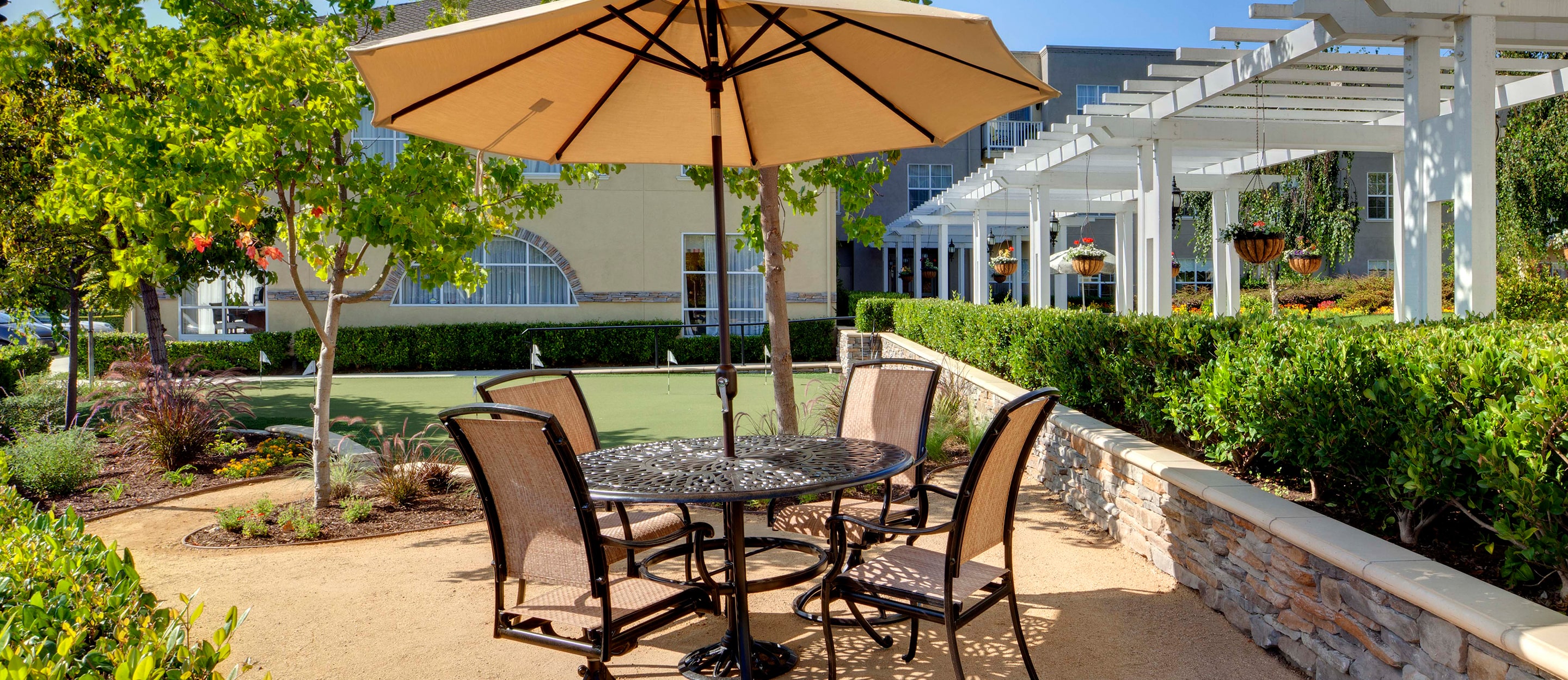 An outdoor patio area with a round metal table and four chairs under a large beige umbrella, surrounded by green landscaping, trees, and a pergola walkway in the background.