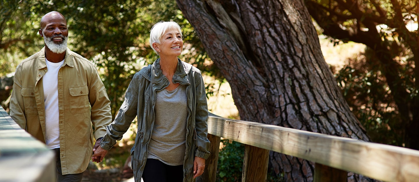 An older couple walks hand in hand on a wooden path in a forest, smiling and enjoying a sunny day surrounded by trees and nature.