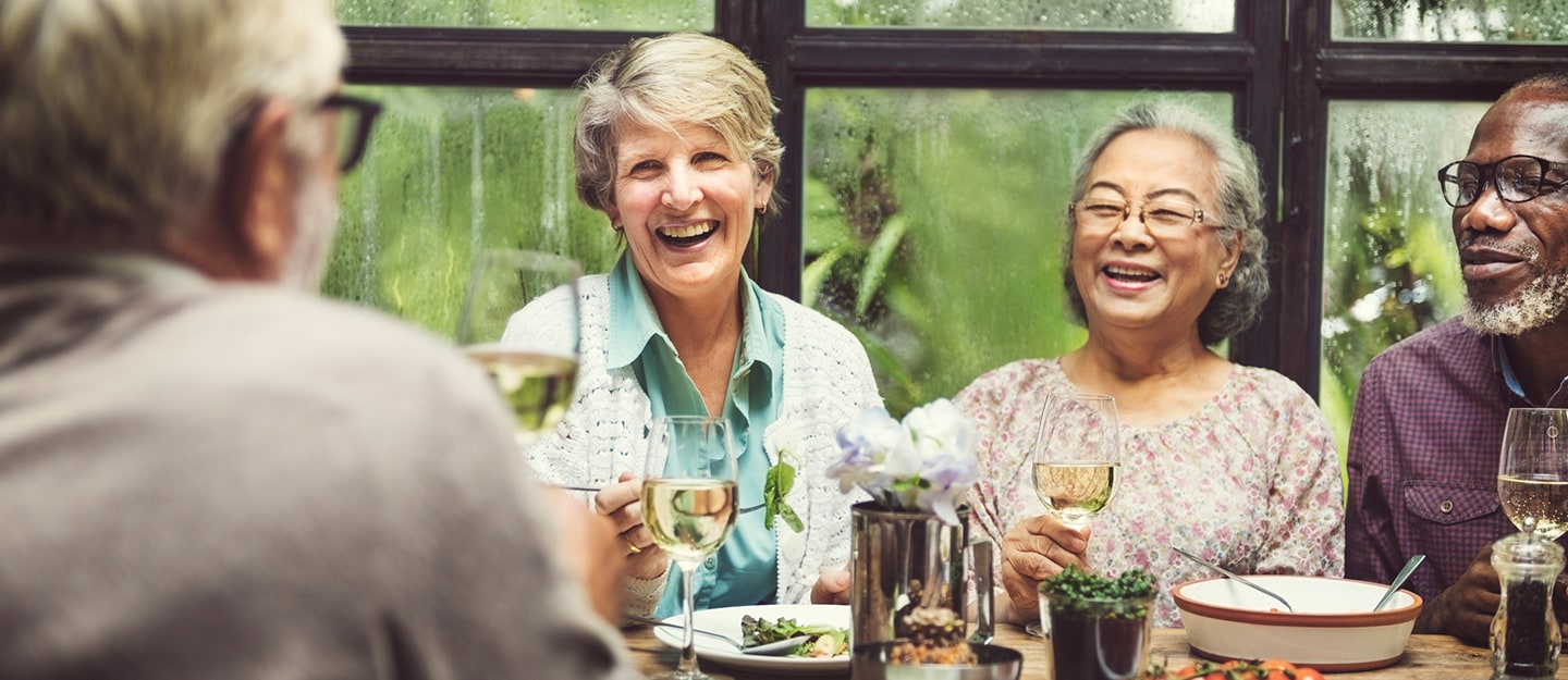 A group of older adults sit around a table, smiling and laughing while enjoying drinks and a meal together in a bright, cozy setting with large windows and greenery outside.
