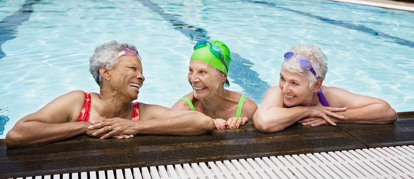Three older women in swimsuits and swim caps smile and laugh together while leaning on the edge of a swimming pool. They appear to be enjoying each others company and the water.