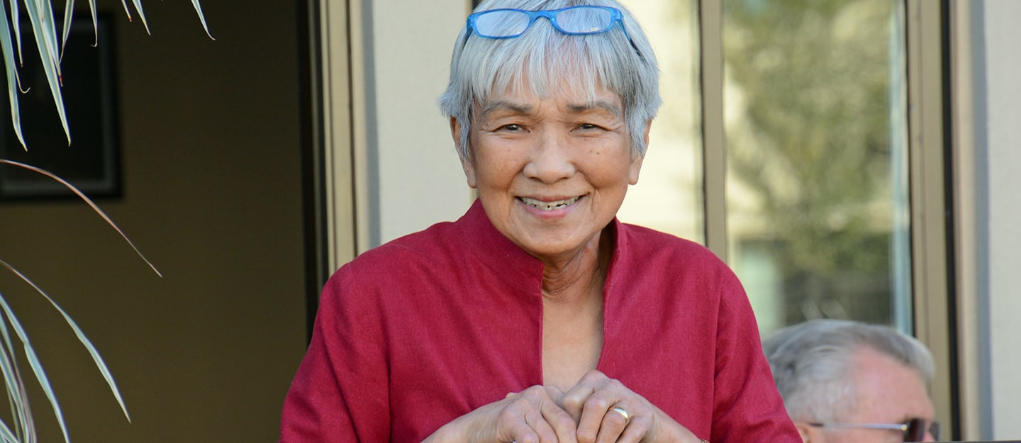 An elderly woman with short gray hair, wearing a red shirt and blue glasses on her head, smiles warmly while standing outdoors near a window.