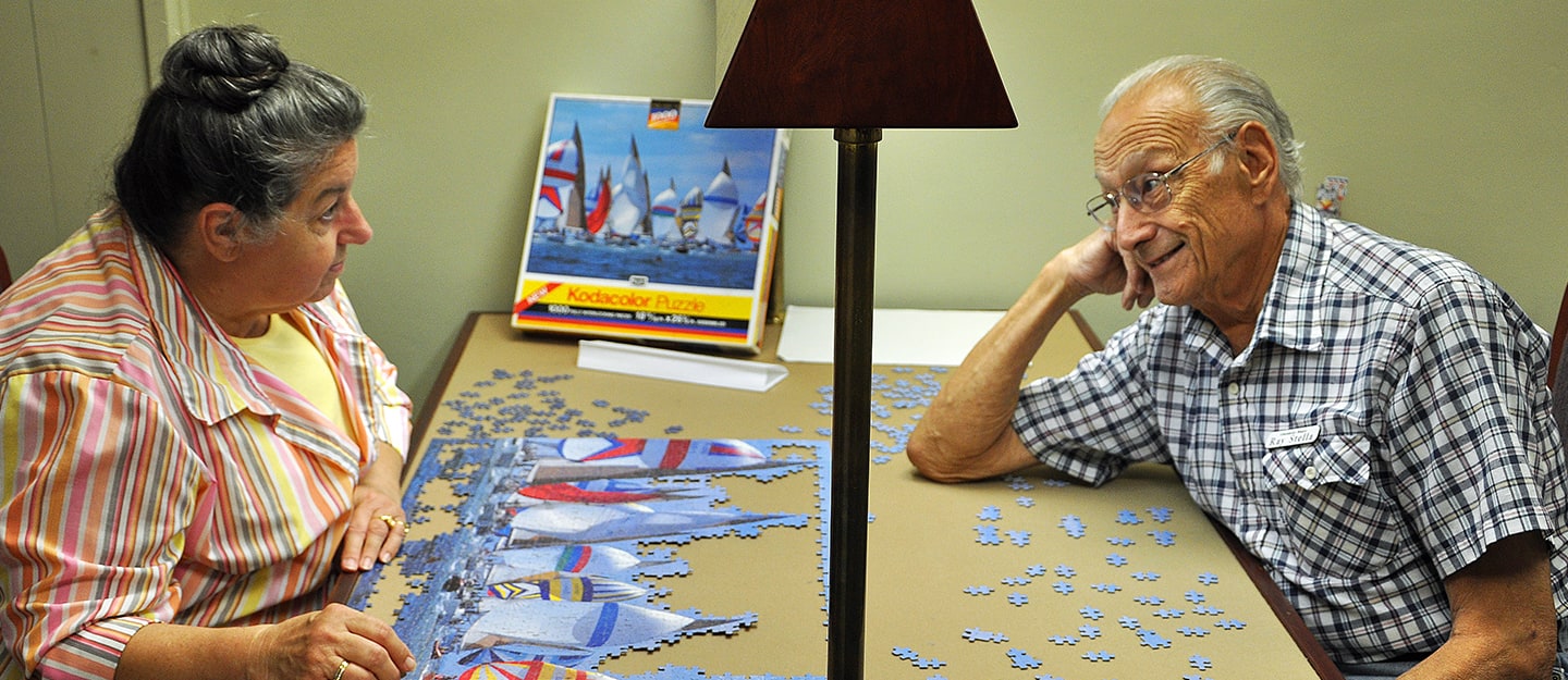 An elderly woman and man sit at a table working on a sailboat jigsaw puzzle together, with the puzzle box visible in the background and a lamp between them.