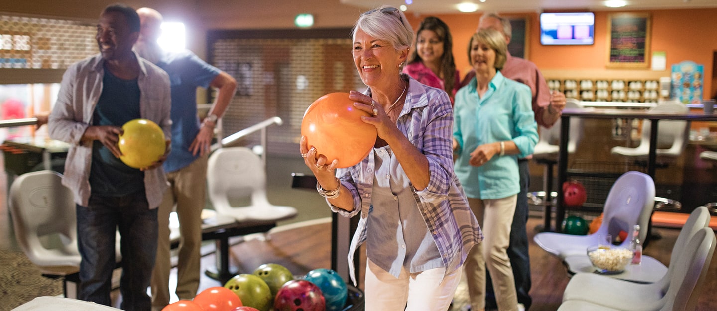 A group of smiling older adults enjoy a game of bowling at an indoor alley; a woman in front holds an orange bowling ball, ready to bowl while her friends cheer her on in the background.