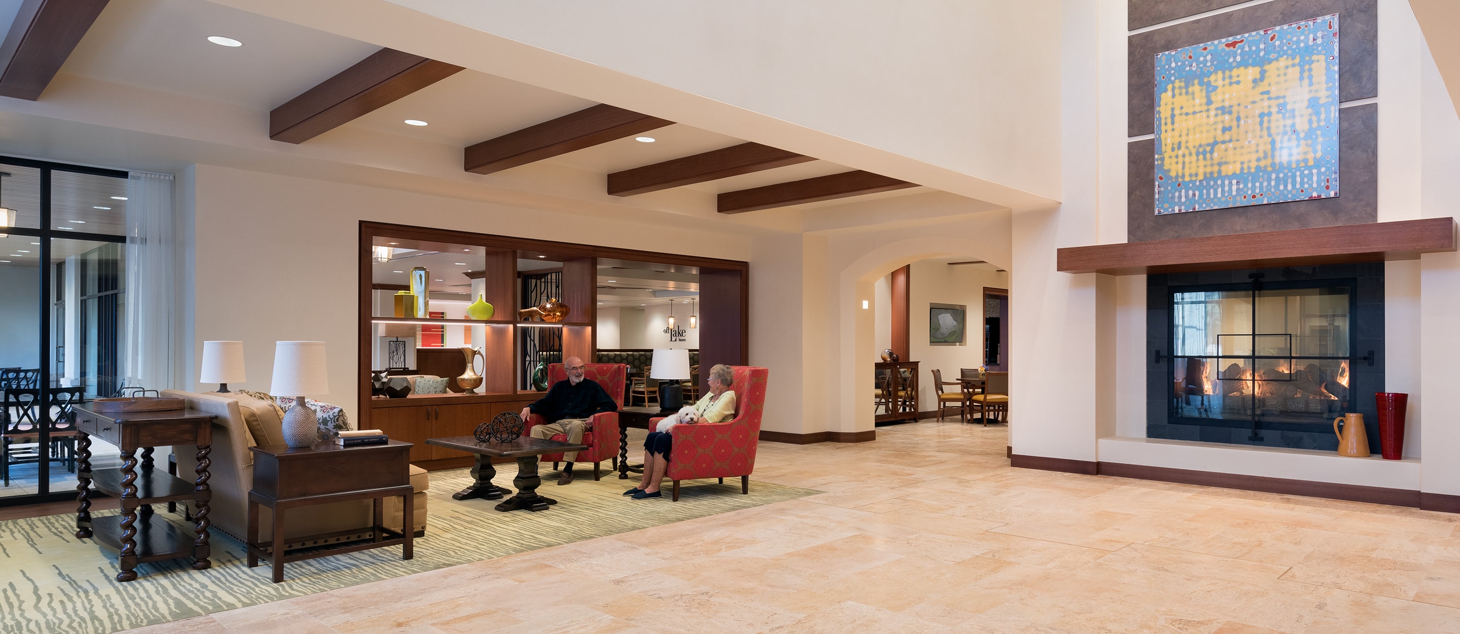 A spacious, modern hotel lobby with tiled floors, a cozy seating area with armchairs, a coffee table, and a fireplace. Large windows and contemporary decor create a warm, inviting atmosphere.