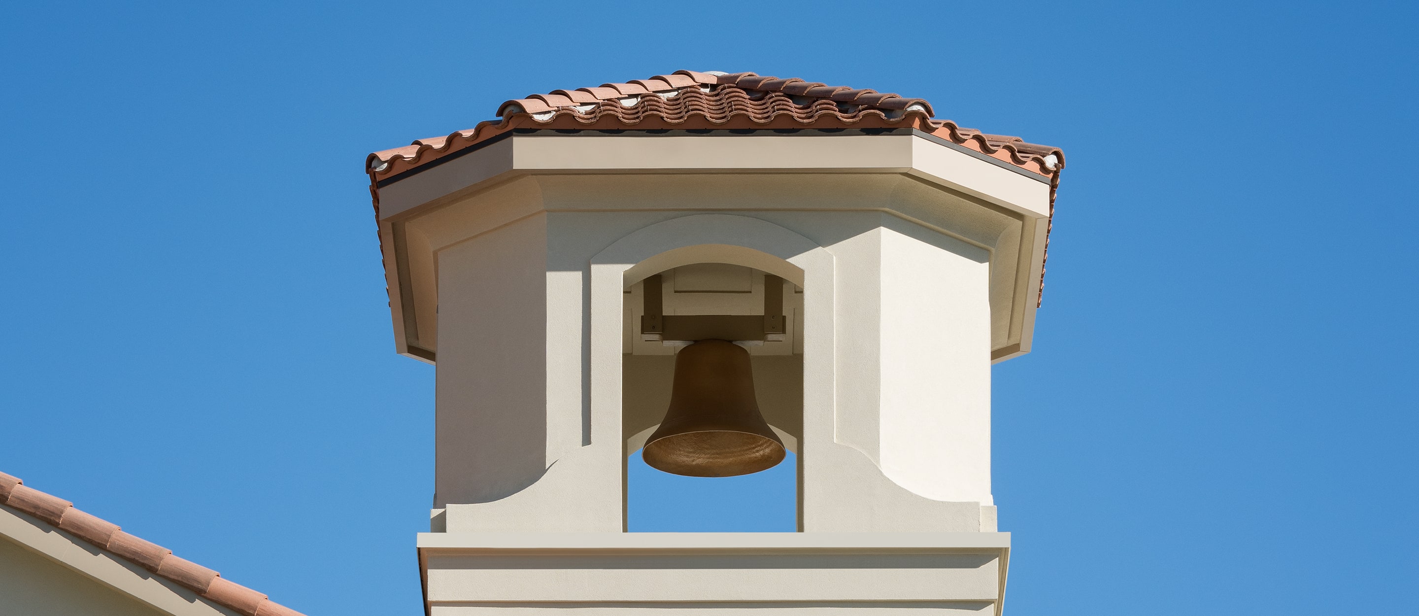 A close-up of a beige bell tower with a red-tiled roof and a bronze bell hanging inside, set against a clear blue sky.