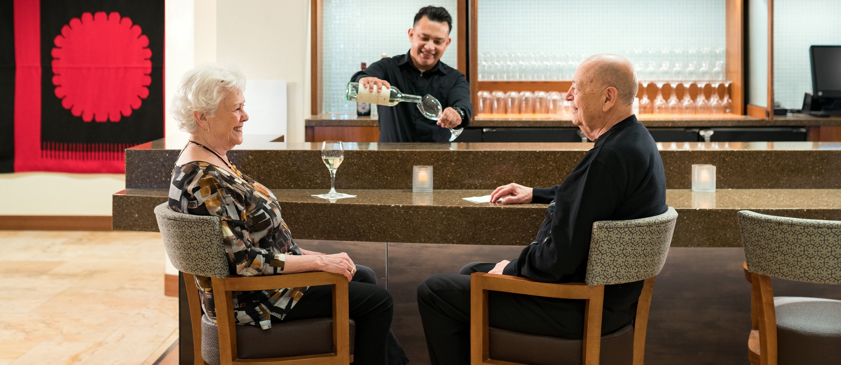 An elderly woman and man sit at a bar, smiling at each other, while a bartender pours white wine into a glass. The bar has modern decor and lit candles on the counter.