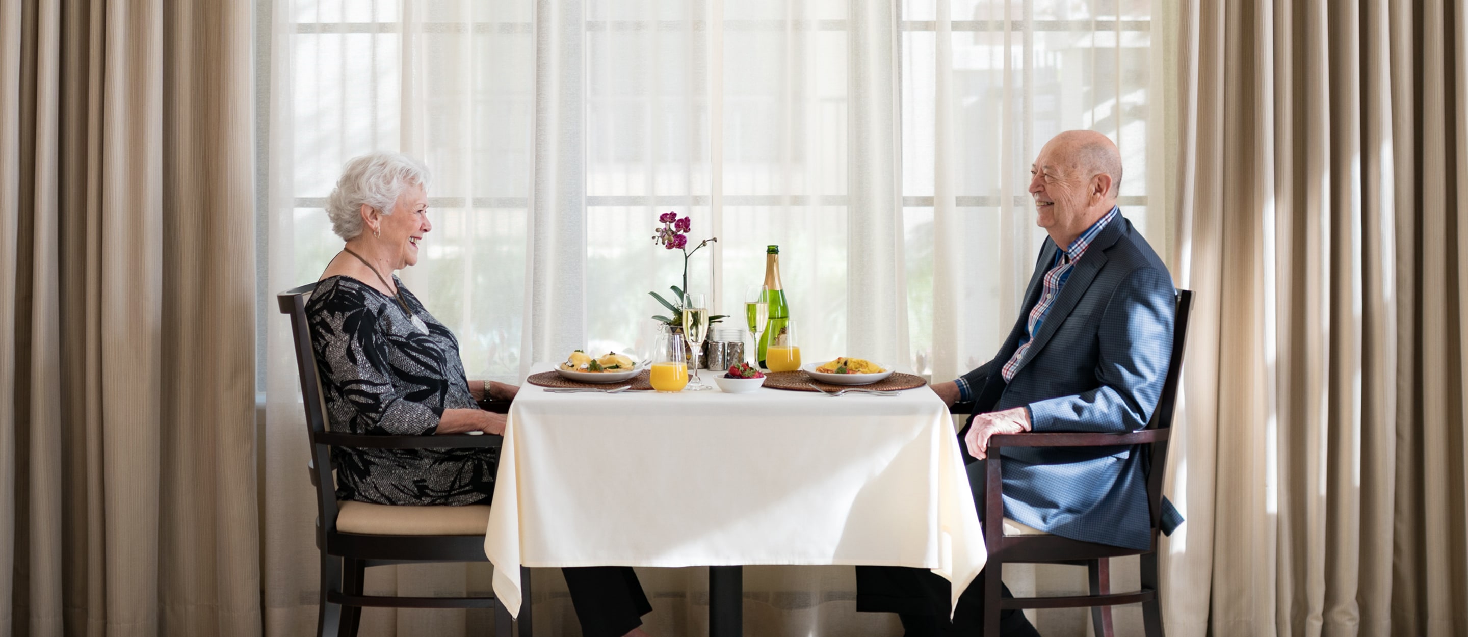 An elderly woman and man sit across from each other at a dining table, smiling and enjoying a meal together. They are dressed formally, and a window with sheer curtains lets in natural light behind them.