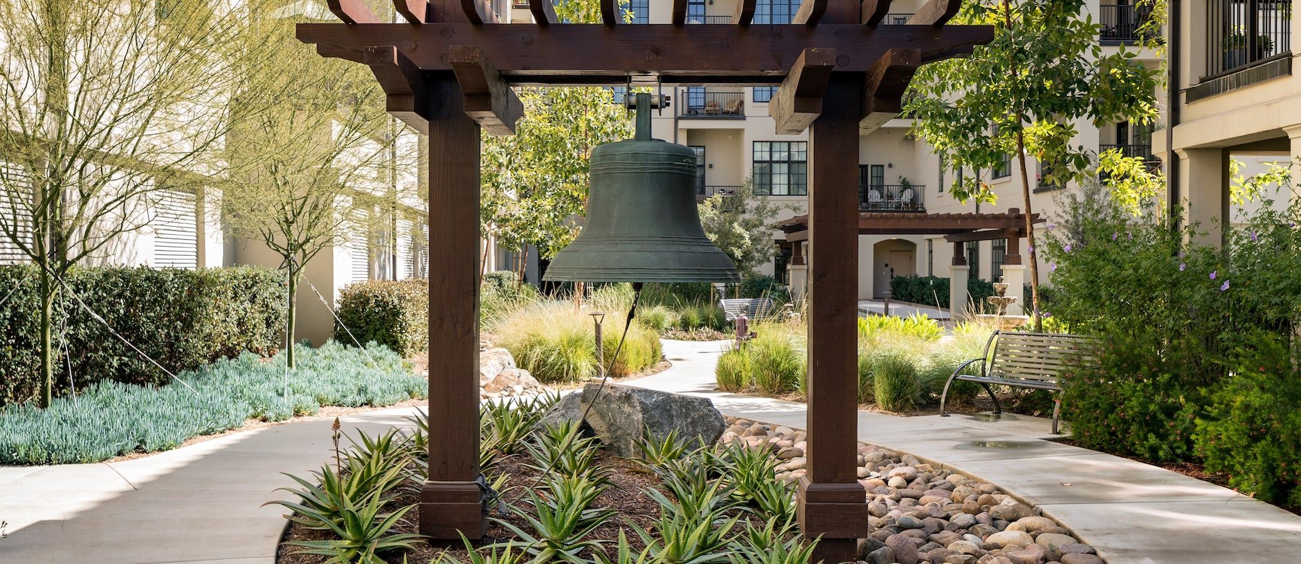 A large bronze bell hangs from a wooden pergola in a landscaped courtyard with winding paths, benches, greenery, and apartment balconies in the background.