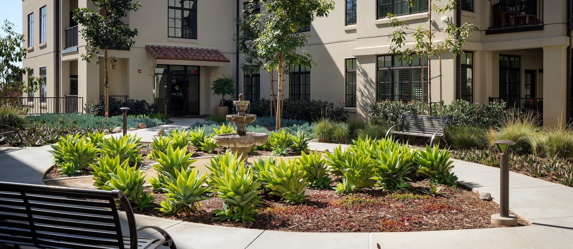 A landscaped courtyard features a circular path, green leafy plants, a stone fountain at the center, benches, and a beige apartment building with large windows in the background.