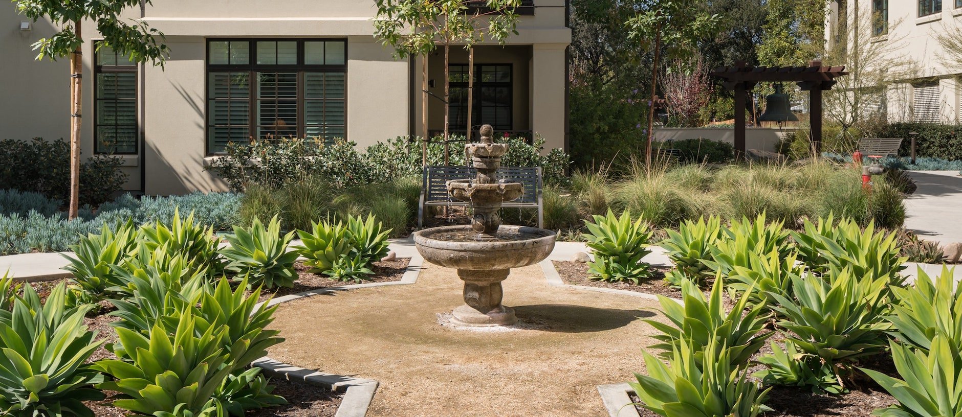 A lush garden courtyard with bright green plants surrounds a stone fountain. A bench sits behind the fountain, and a beige building with shuttered windows is in the background. Sunlight casts soft shadows.