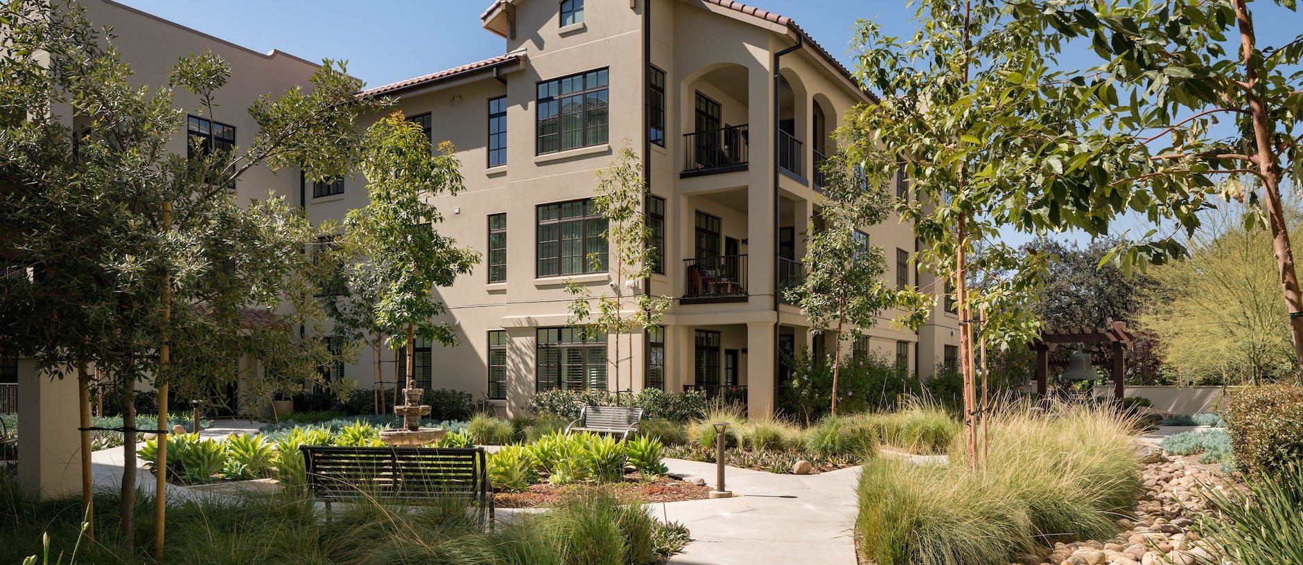 Three-story beige apartment building with balconies, surrounded by lush greenery, trees, benches, a small fountain, and a winding walkway under a clear blue sky.