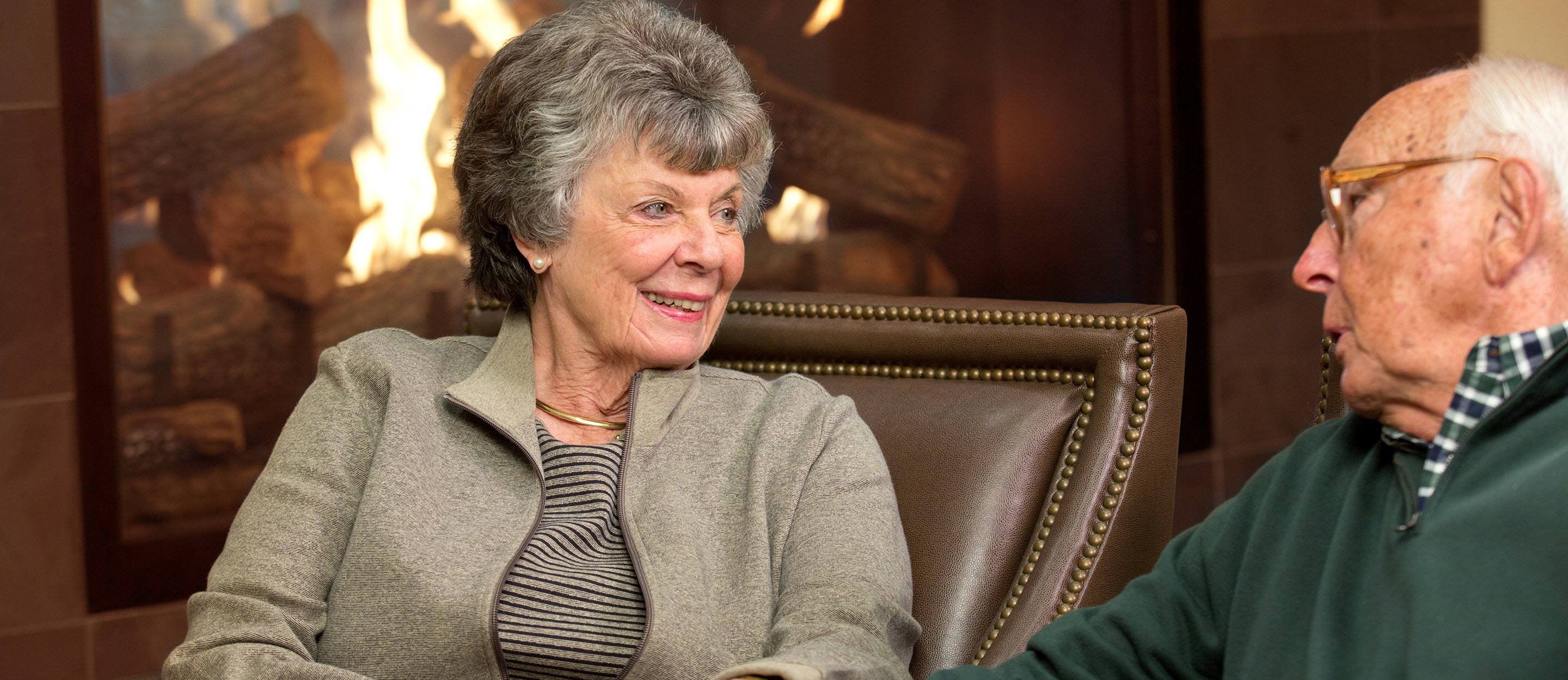 An older woman and man sit together in armchairs, smiling and talking in front of a lit fireplace, creating a warm and cozy atmosphere.
