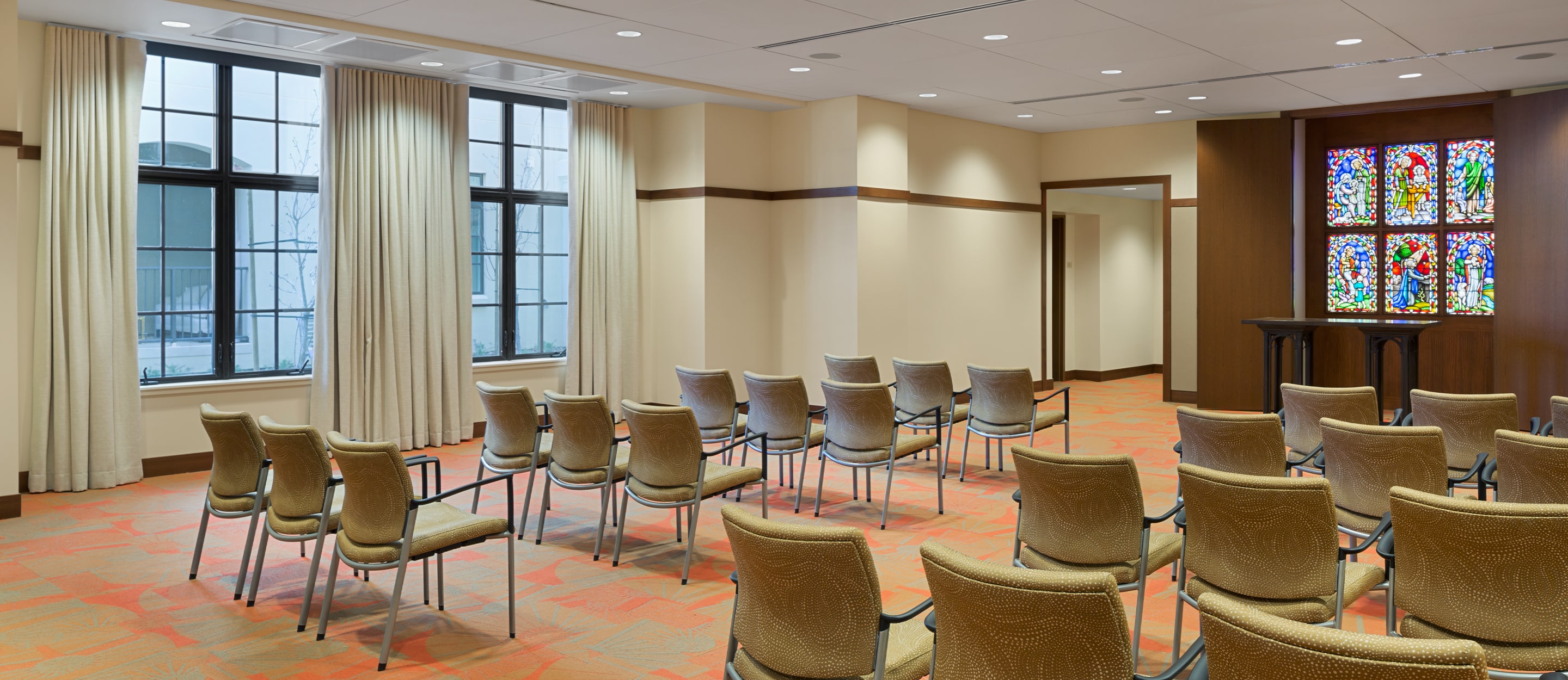 A small room with rows of beige chairs facing a stained glass window and podium. Large windows with cream curtains line one wall, and the space is softly lit with recessed ceiling lights.