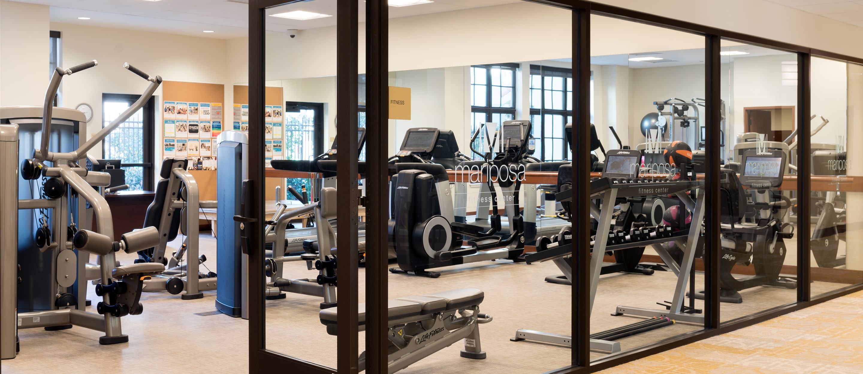 A modern gym with large windows, filled with various exercise equipment including treadmills, ellipticals, weight machines, and benches. The space is clean, well-lit, and has light-colored flooring.