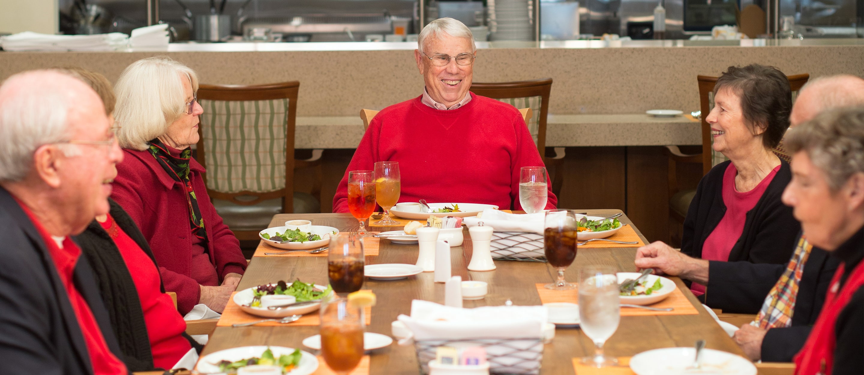A group of six older adults sit around a dining table, smiling and talking while enjoying a meal together in a bright, casual restaurant setting.