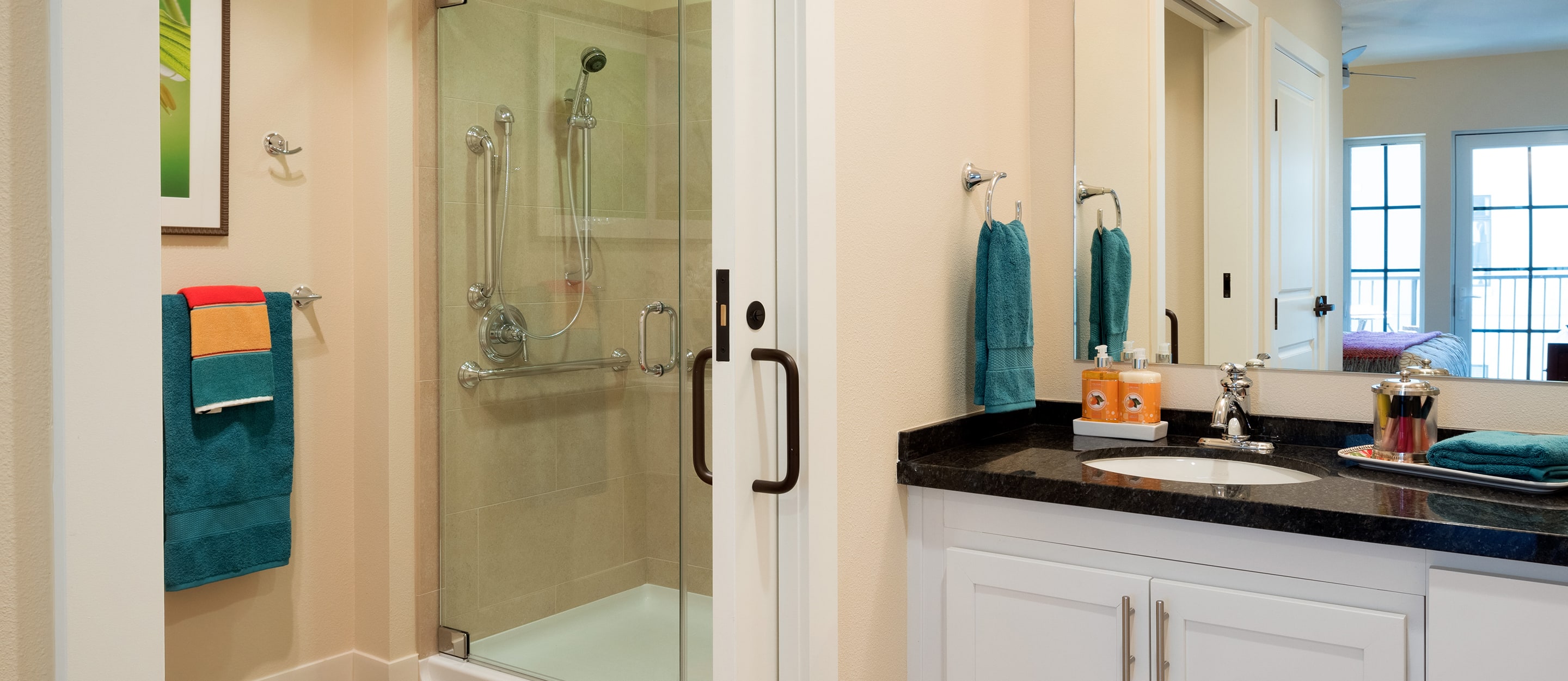 Modern bathroom with a glass-enclosed shower, white vanity with a black countertop, sink, teal towels, and toiletries. A colorful towel hangs on the wall next to the shower. Large mirrors and natural light brighten the space.