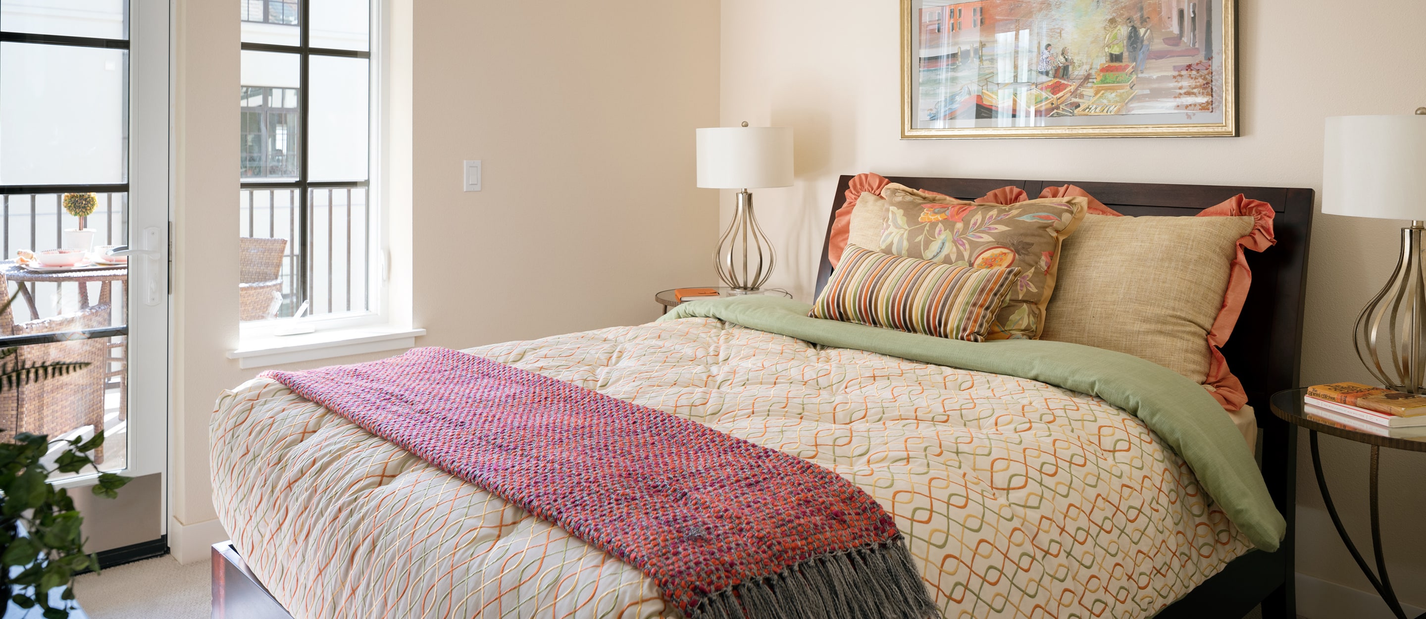 A cozy bedroom with a neatly made bed featuring patterned bedding, throw pillows, and a purple throw blanket. Two bedside tables with lamps, a painting above the bed, and a door open to a sunny outdoor patio.