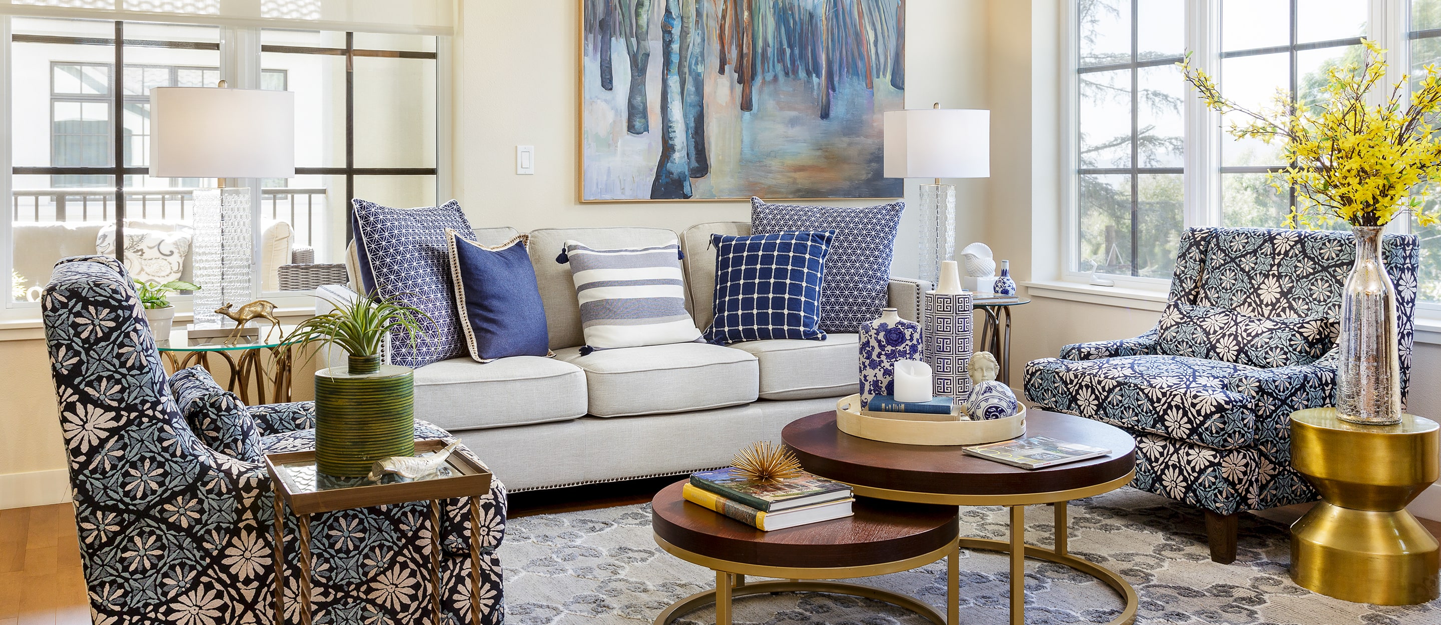 Stylish living room with a cream sofa, patterned blue and white chairs, blue accent pillows, two round wooden coffee tables with books and decor, table lamps, and large artwork above the sofa. Large windows bring in natural light.
