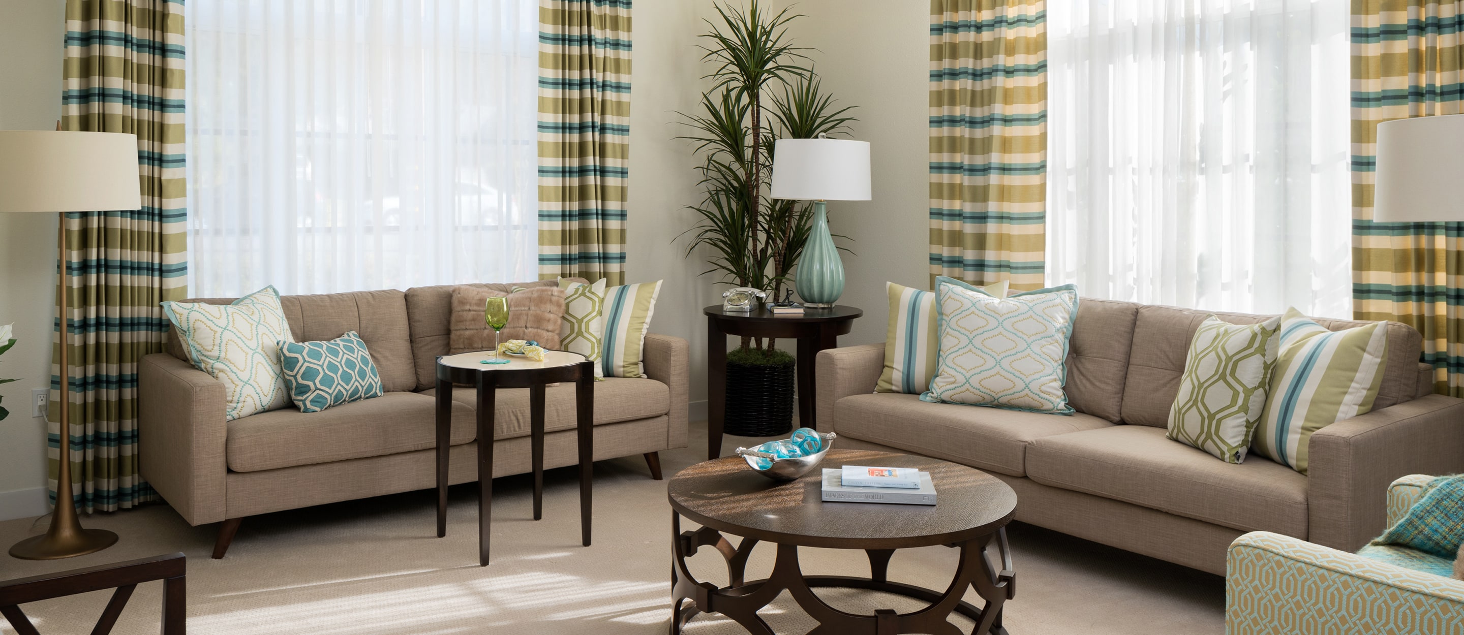 A bright, modern living room with two beige sofas, colorful patterned pillows, striped curtains, tall potted plants, a round wooden coffee table, and large windows letting in natural light.