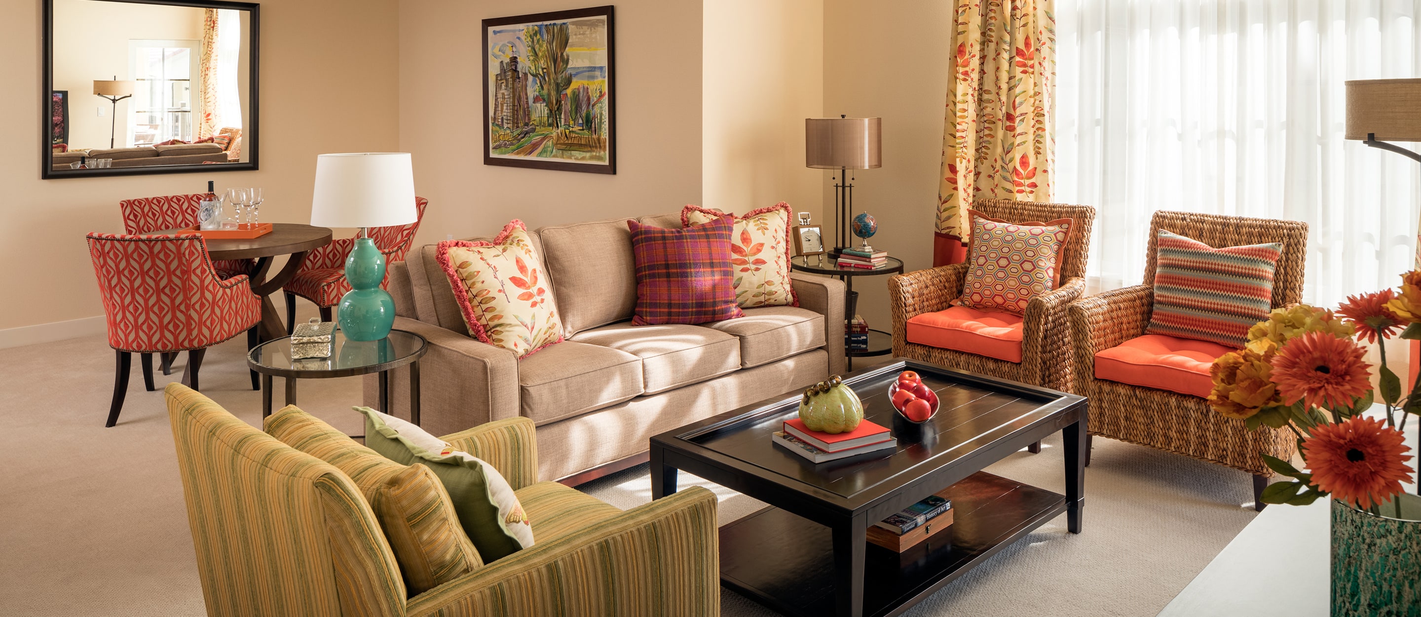 A cozy living room with a beige sofa, two wicker armchairs with orange cushions, a striped green chair, a black coffee table, colorful pillows, floral curtains, and a round dining table with red chairs in the background.