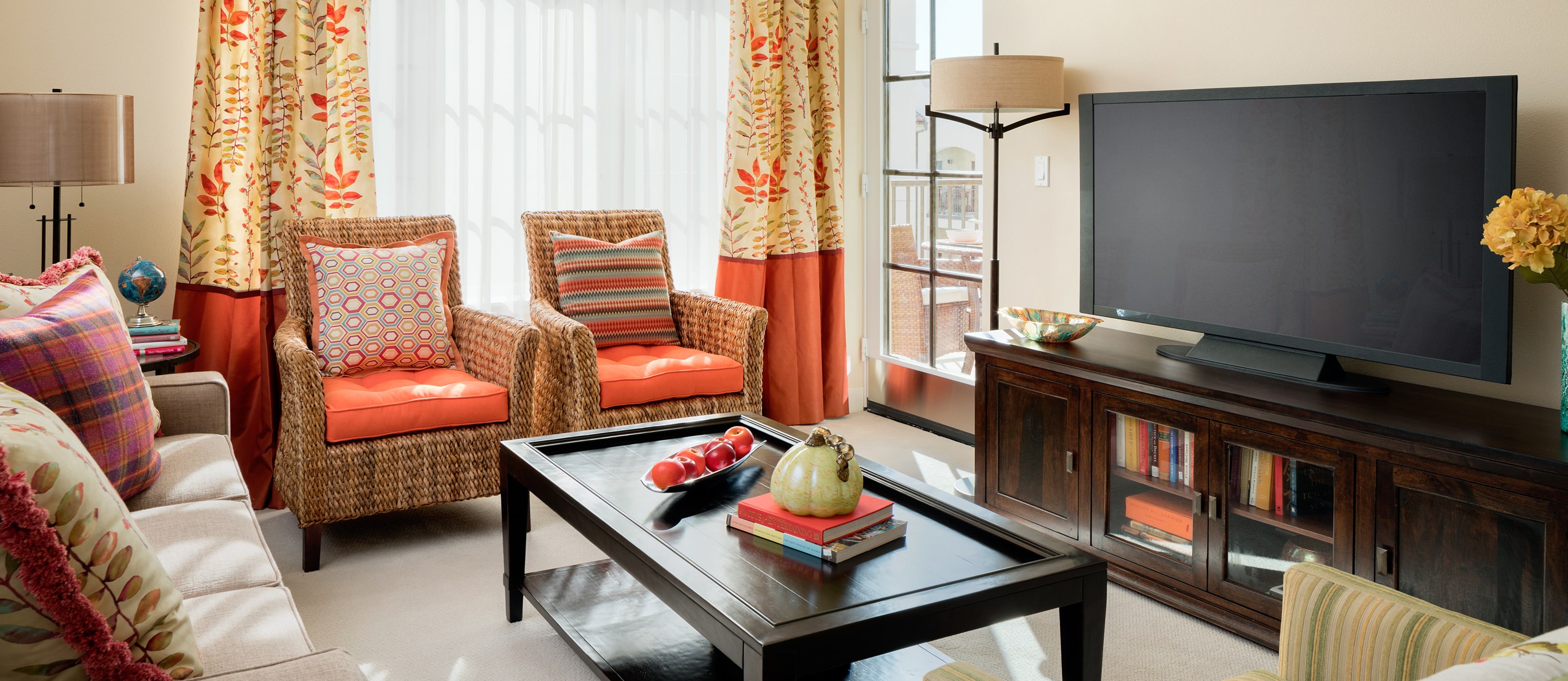 A bright living room with wicker chairs, a beige sofa with colorful pillows, a dark wood coffee table, a TV on a wooden stand, and large windows with patterned curtains letting in natural light.