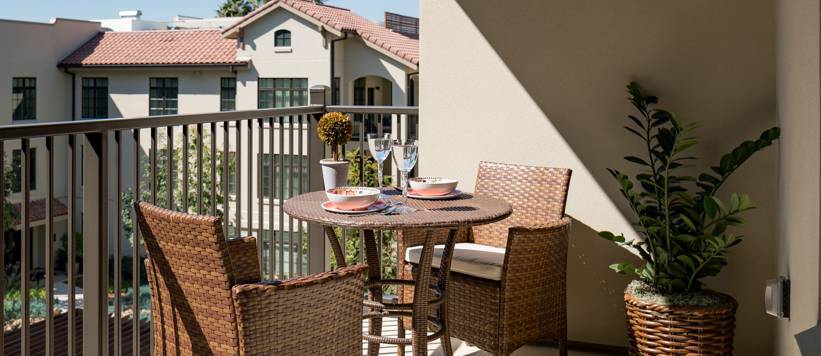 A small balcony with a round wicker table set for two, featuring bowls, plates, and wine glasses. Two wicker chairs and a potted plant are nearby, overlooking a building with red-tiled roofs.