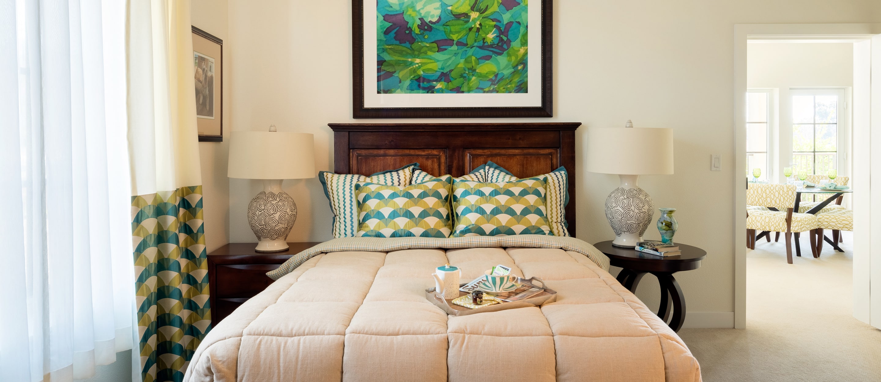 A cozy bedroom with a beige comforter, patterned pillows, and a dark wood headboard. Two lamps sit on bedside tables. Above the bed hangs a colorful abstract painting. A tray with a cup and books rests on the bed.