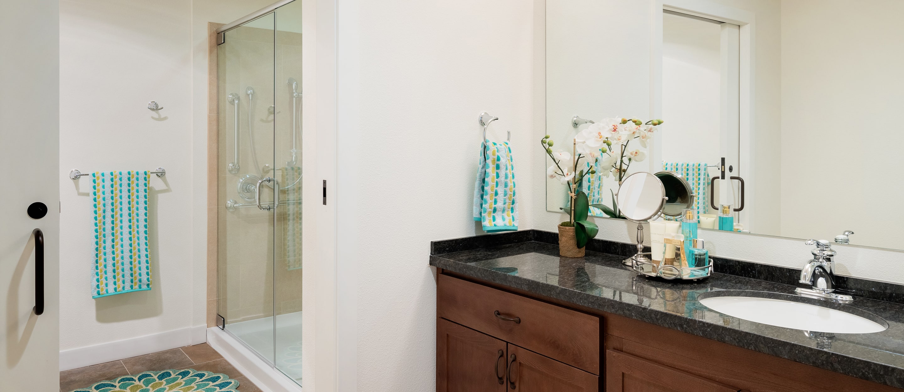Modern bathroom with a glass-enclosed shower, brown vanity with a black countertop, sink, mirror, potted orchid, and teal-patterned towels and rug accents.